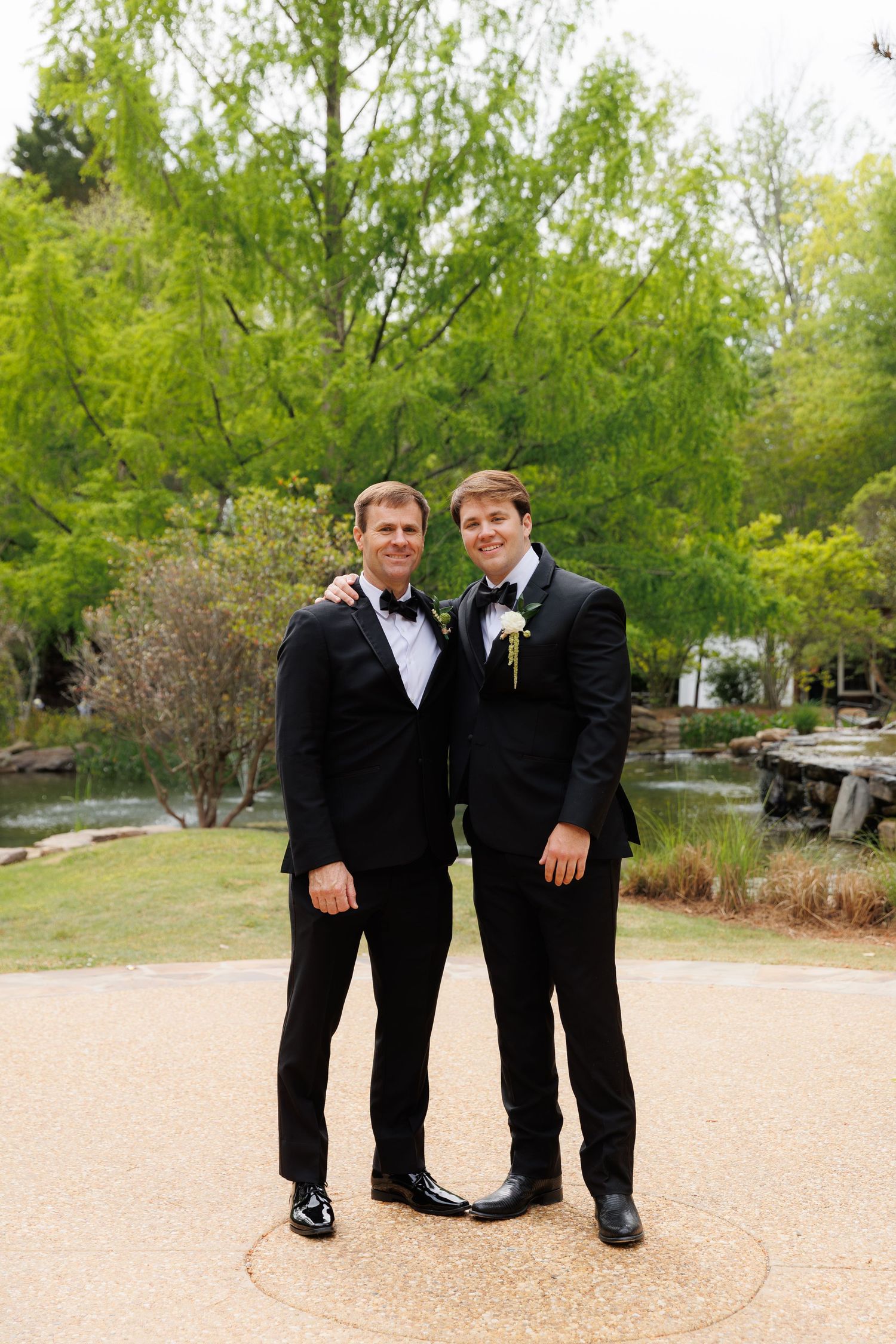 Two men in black tuxedos with boutonnieres pose together smiling in an outdoor garden wedding setting.