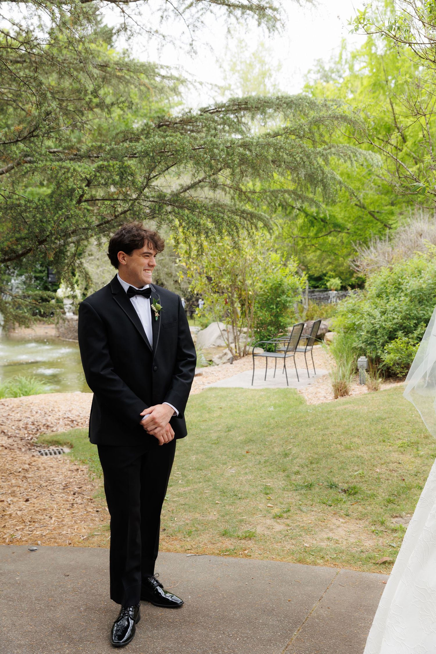 Groom in black tuxedo with bow tie smiles while waiting for bride during outdoor garden first look.