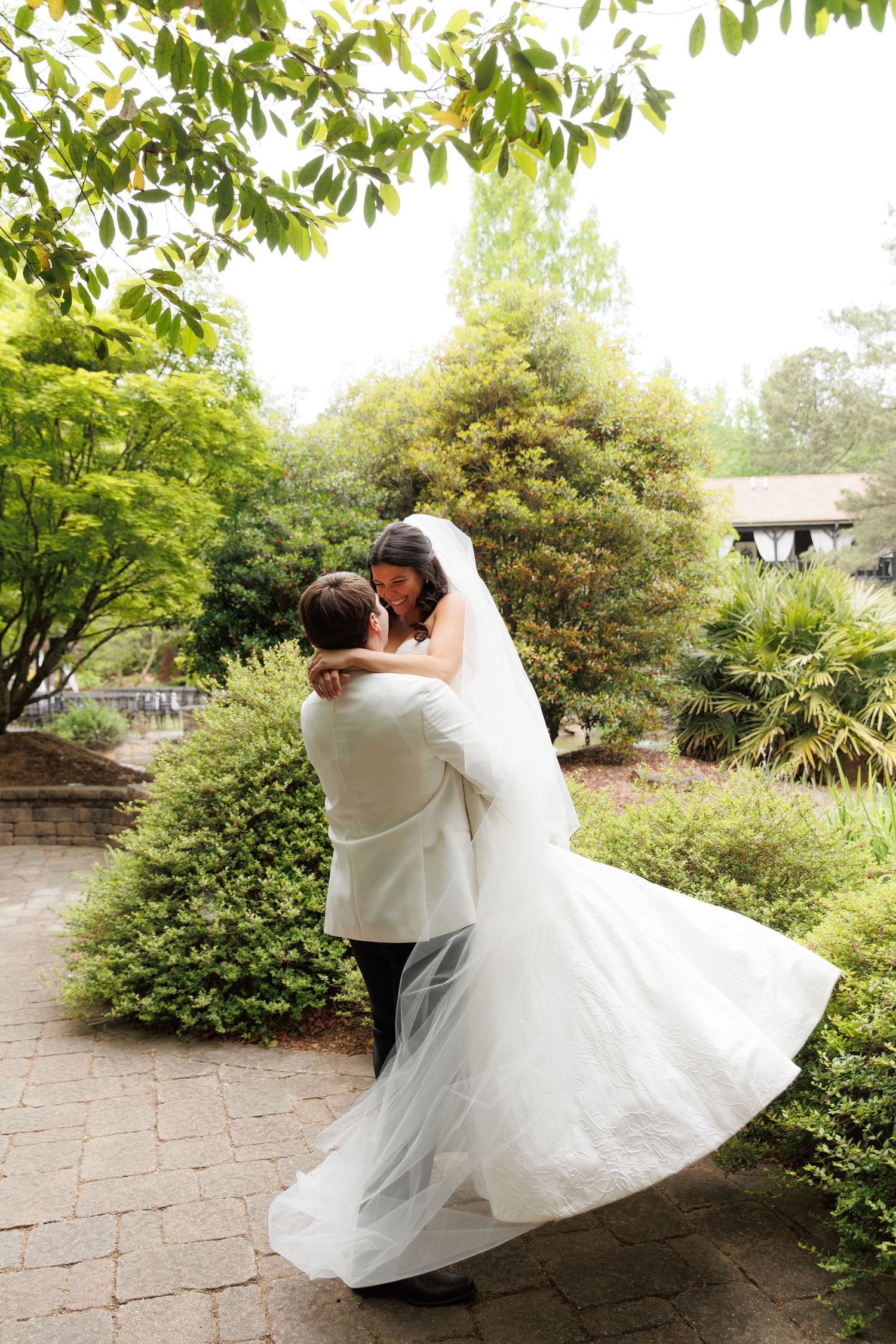Groom lifting bride in white gown, sharing romantic moment in lush garden setting on wedding day.