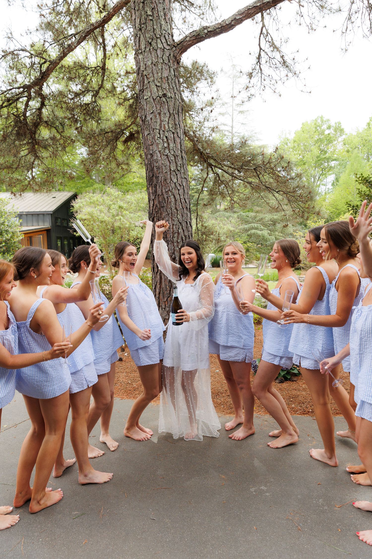 Bride in white robe celebrates with bridesmaids in matching blue pajamas outdoors, popping champagne under pine trees.