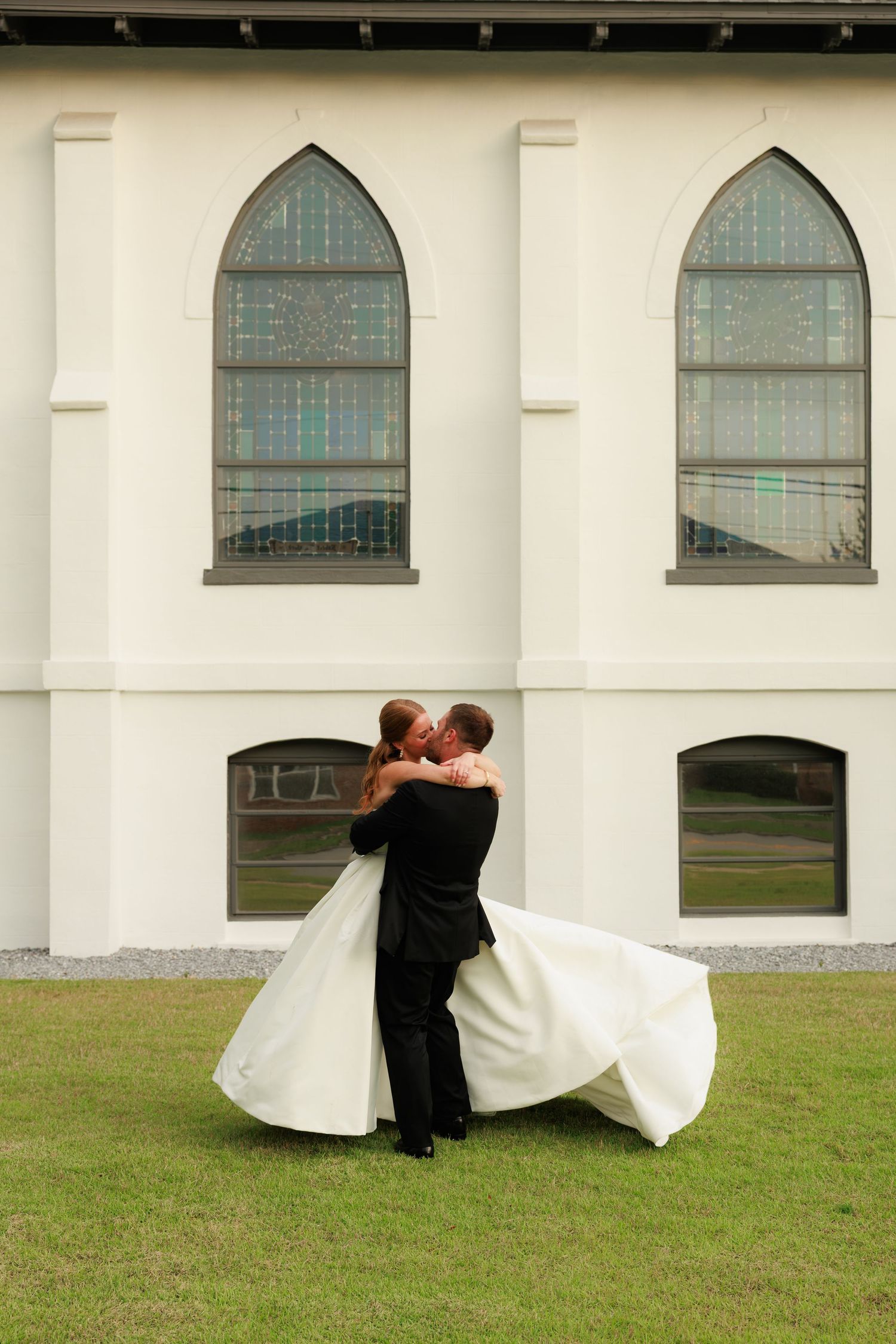 Groom lifts bride in romantic embrace on church lawn, her white ball gown flowing in the breeze.
