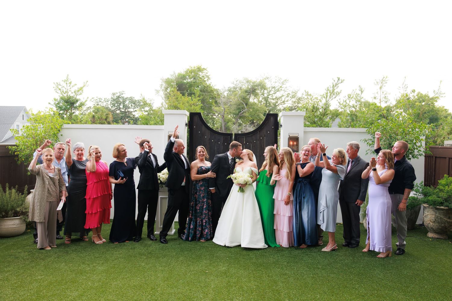 Wedding party group photo with bride and groom kissing surrounded by cheering guests on green lawn.