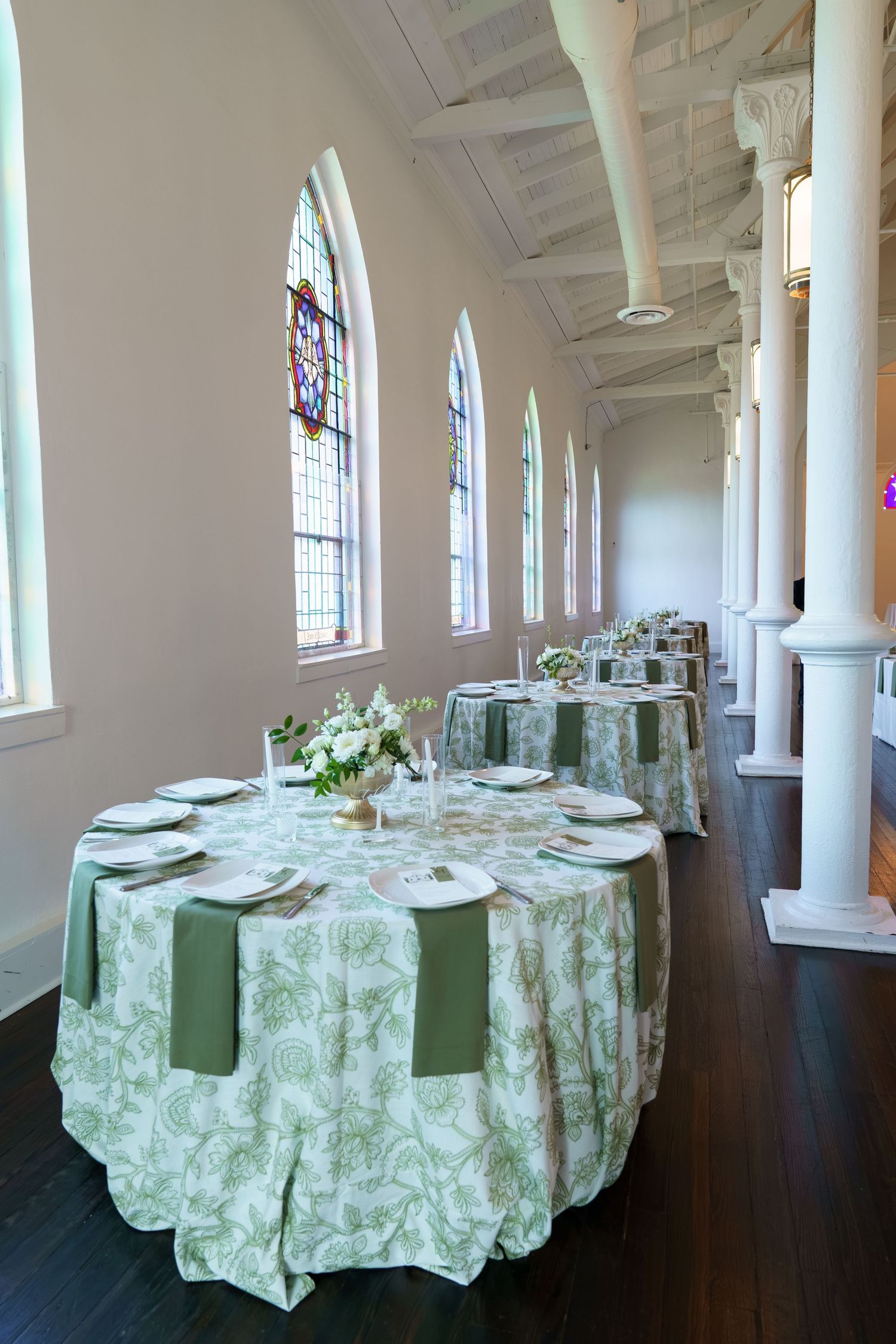 Elegant wedding reception tables with green floral linens and white chairs in a historic venue with stained glass windows.