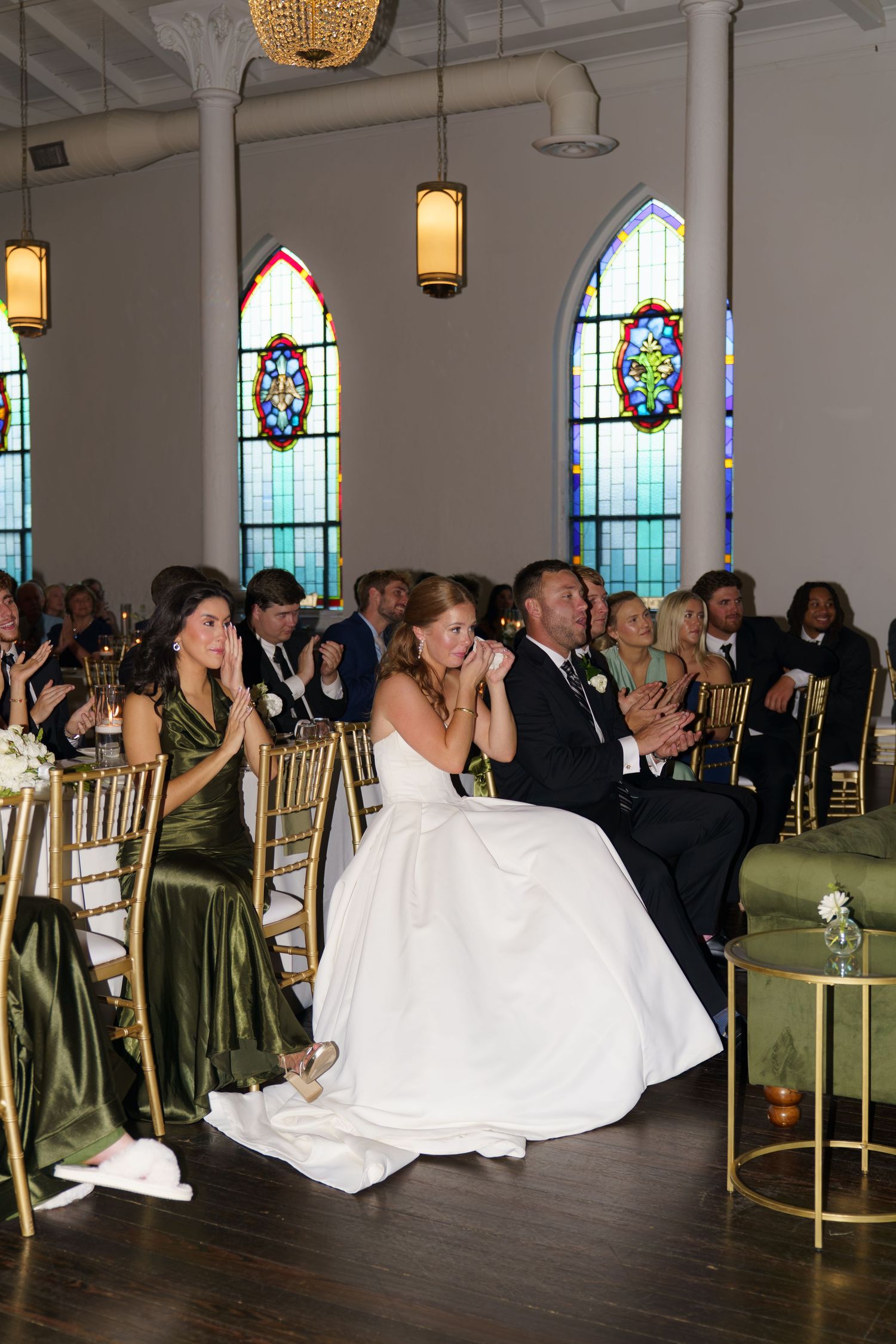 Bride and groom clap joyfully while seated at wedding reception in a beautiful venue with stained glass windows.
