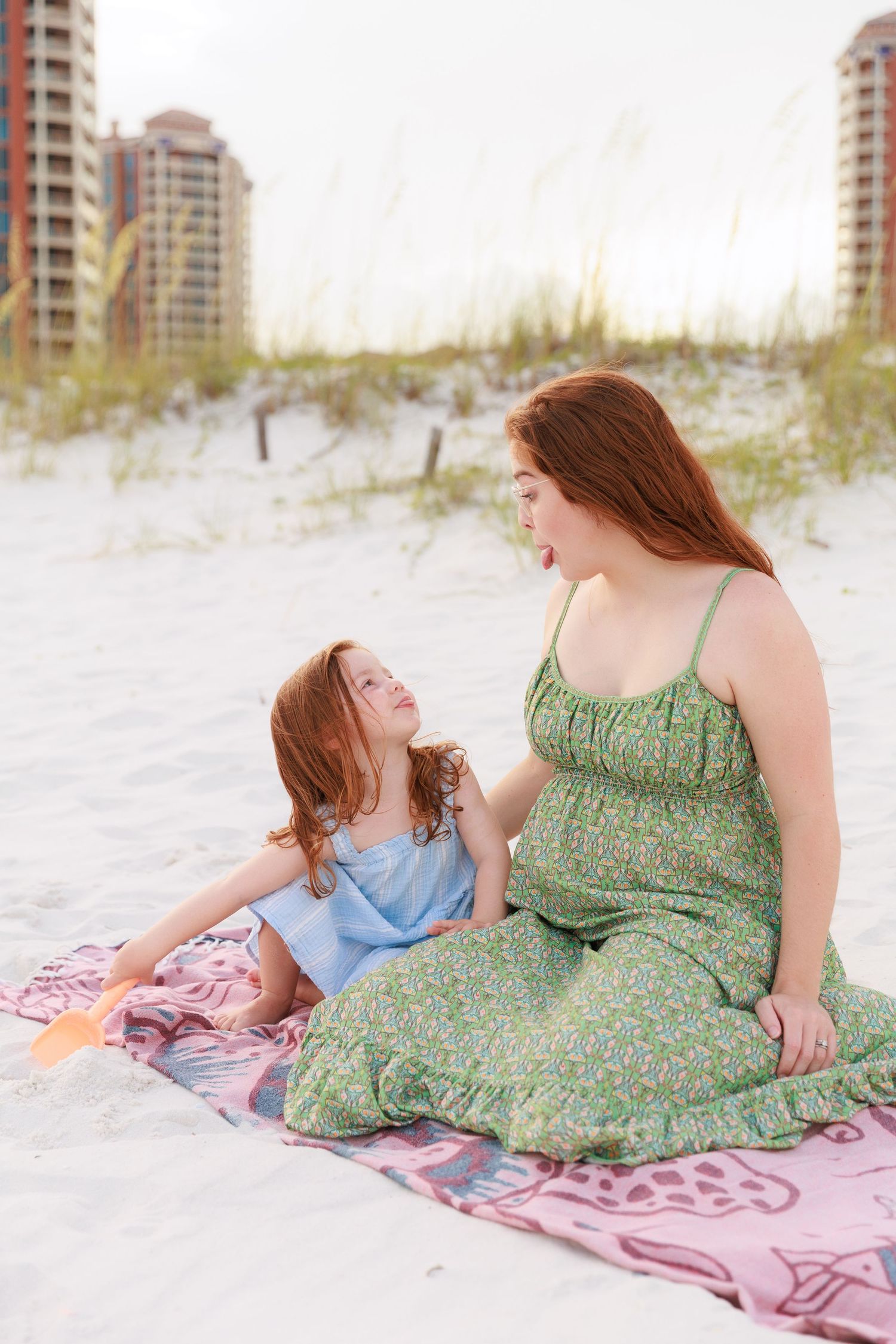 Two people relaxing on a pink blanket on white sand beach with beachfront condos visible.