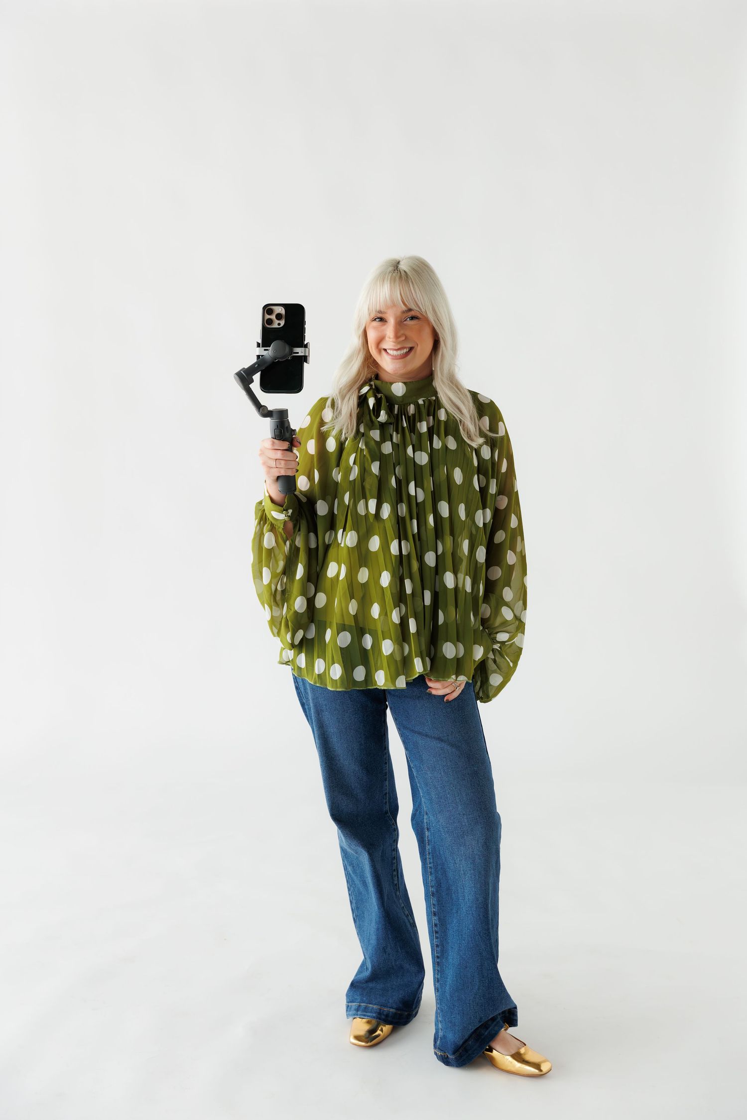 Someone wearing a green polka dot blouse and jeans holds a camera gimbal while standing against a white background.
