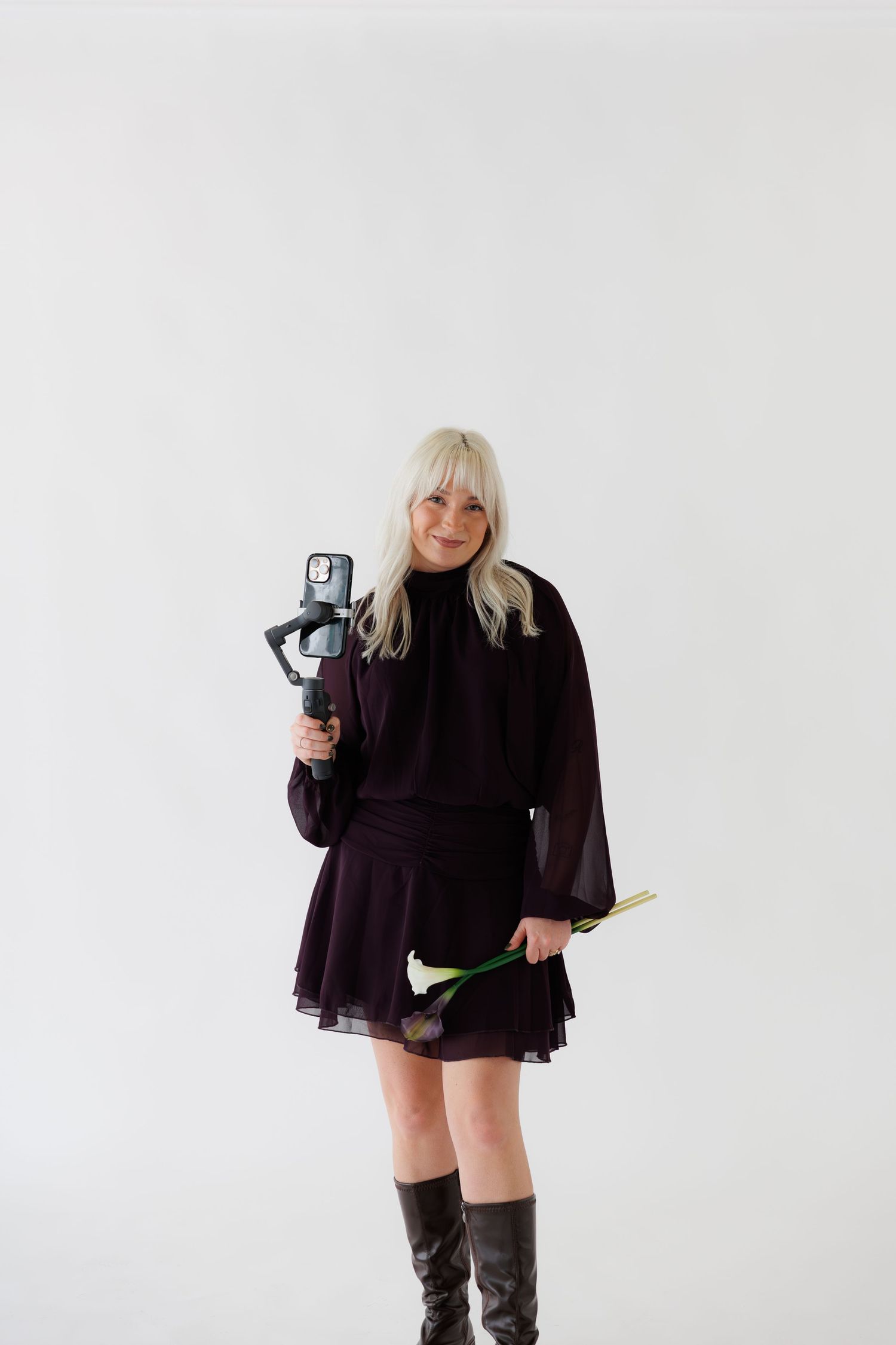 Content creator filming with handheld camera while posing in dark outfit and boots.