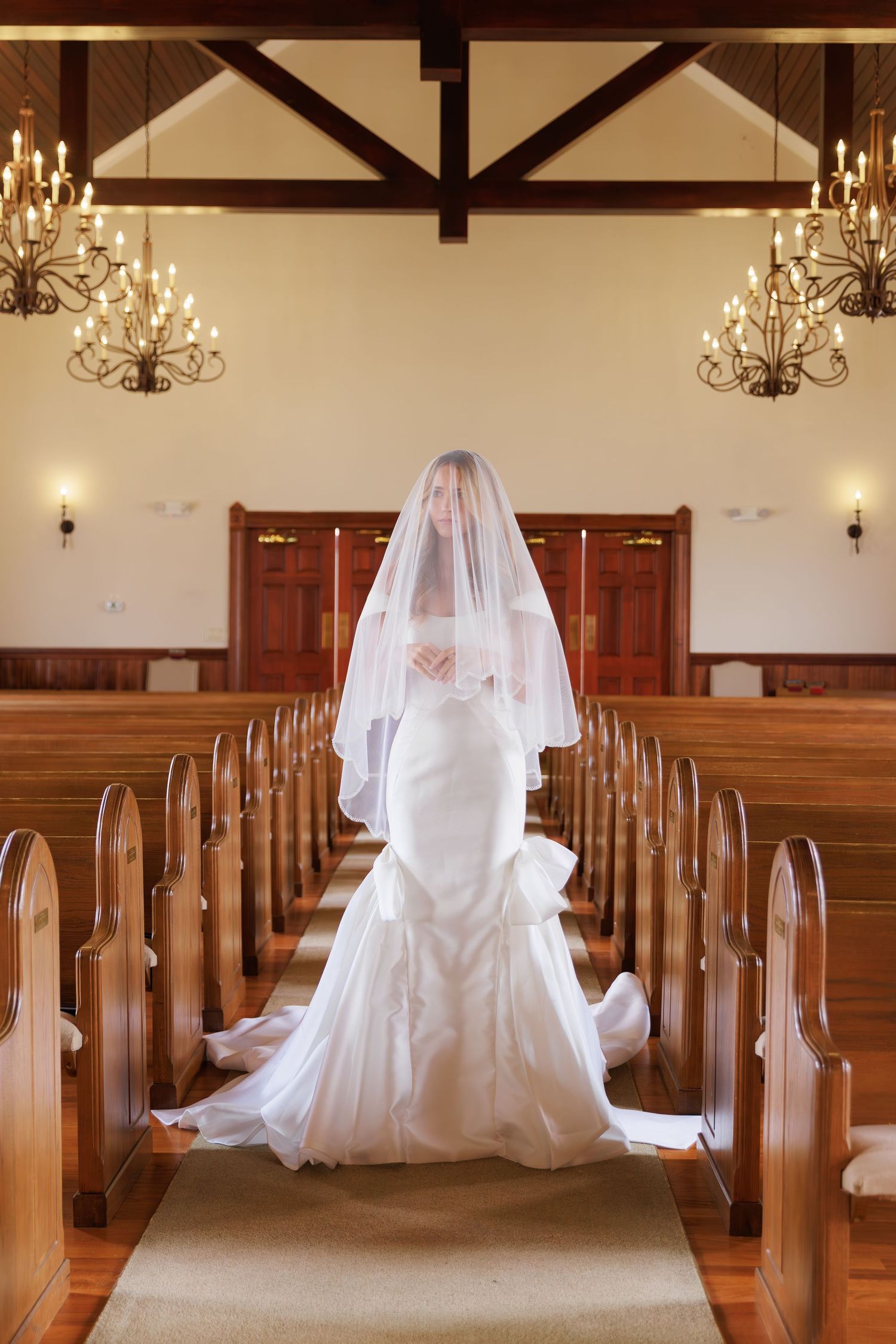 Elegant white bridal gown photographed in sequence along a church aisle with wooden pews and chandeliers above.