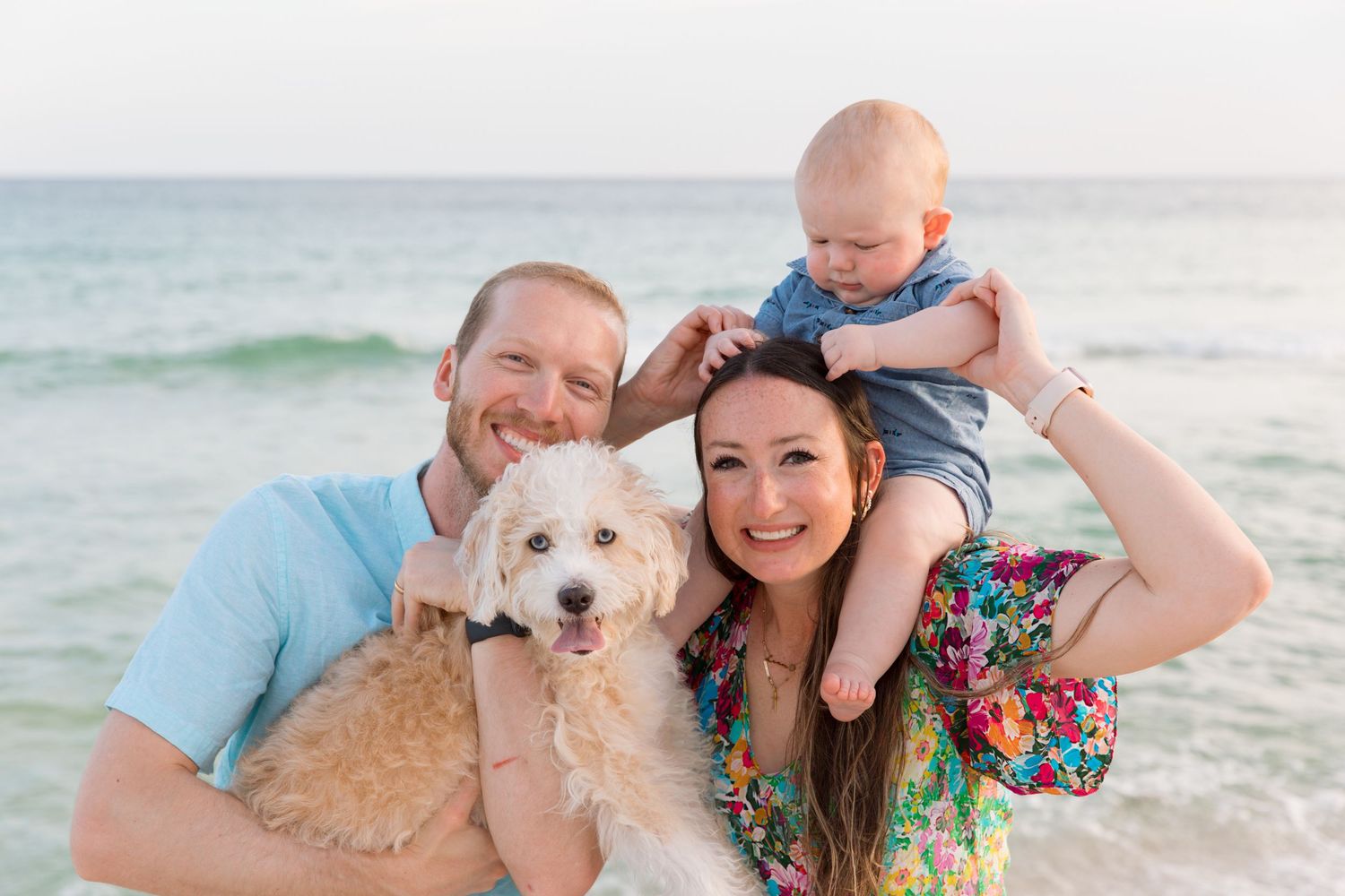 Couple with their dog enjoying a sunny day at the beach with waves and sand in the background.