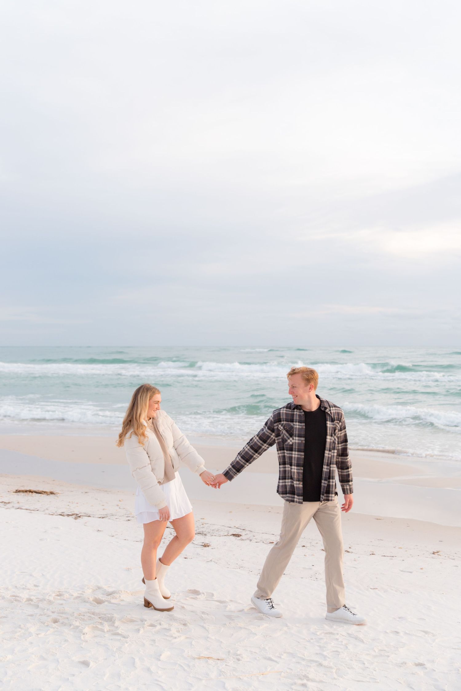 Couple holding hands while walking and playing on white sandy beach with ocean waves.