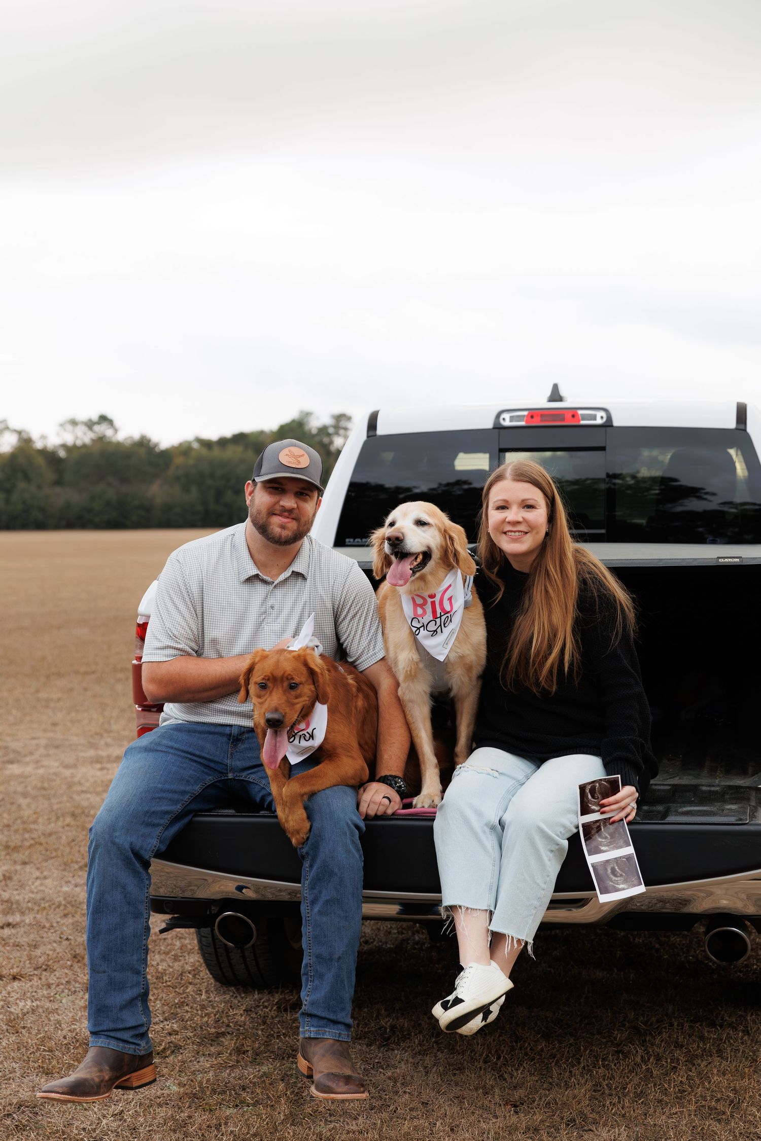 A couple sits on a black pickup truck tailgate with their two dogs during a countryside photoshoot.