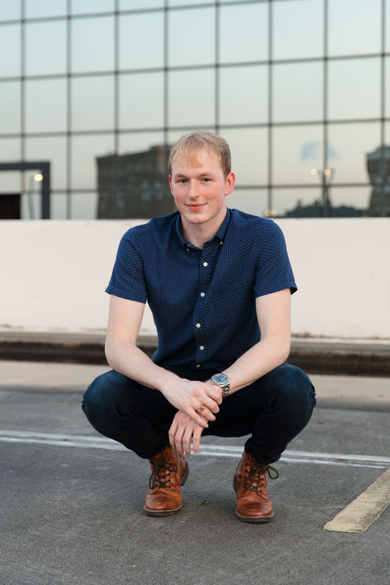 Young man in navy polo shirt squatting outdoors near glass building, wearing brown boots and a watch.