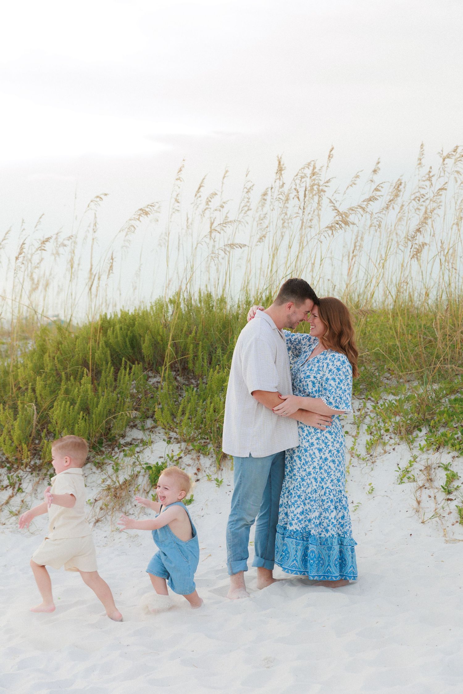 A couple shares a romantic moment on white sand beach dunes while two toddlers play nearby amid sea oats.