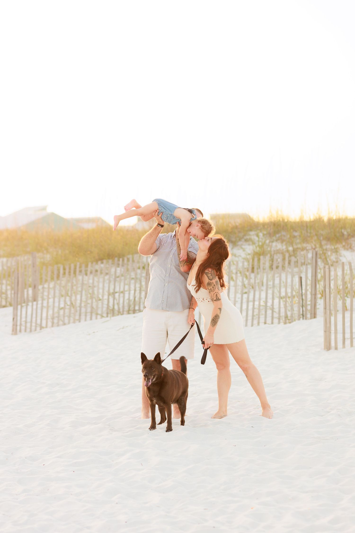 Father lifting toddler for a kiss while mother watches and dog stands on white sand beach.
