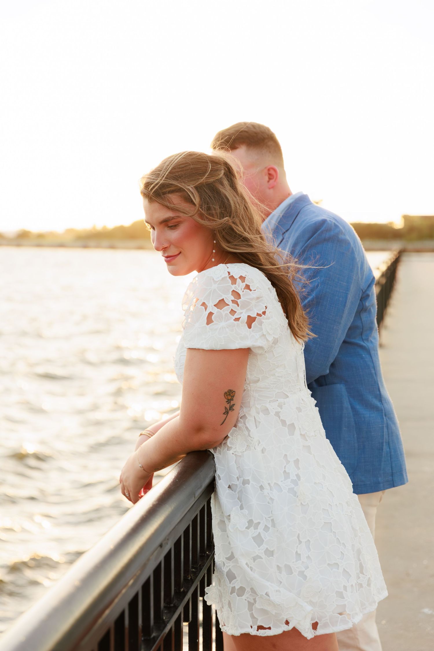 Woman in white lace dress leaning on a railing at golden hour waterfront, man in blue blazer behind her.