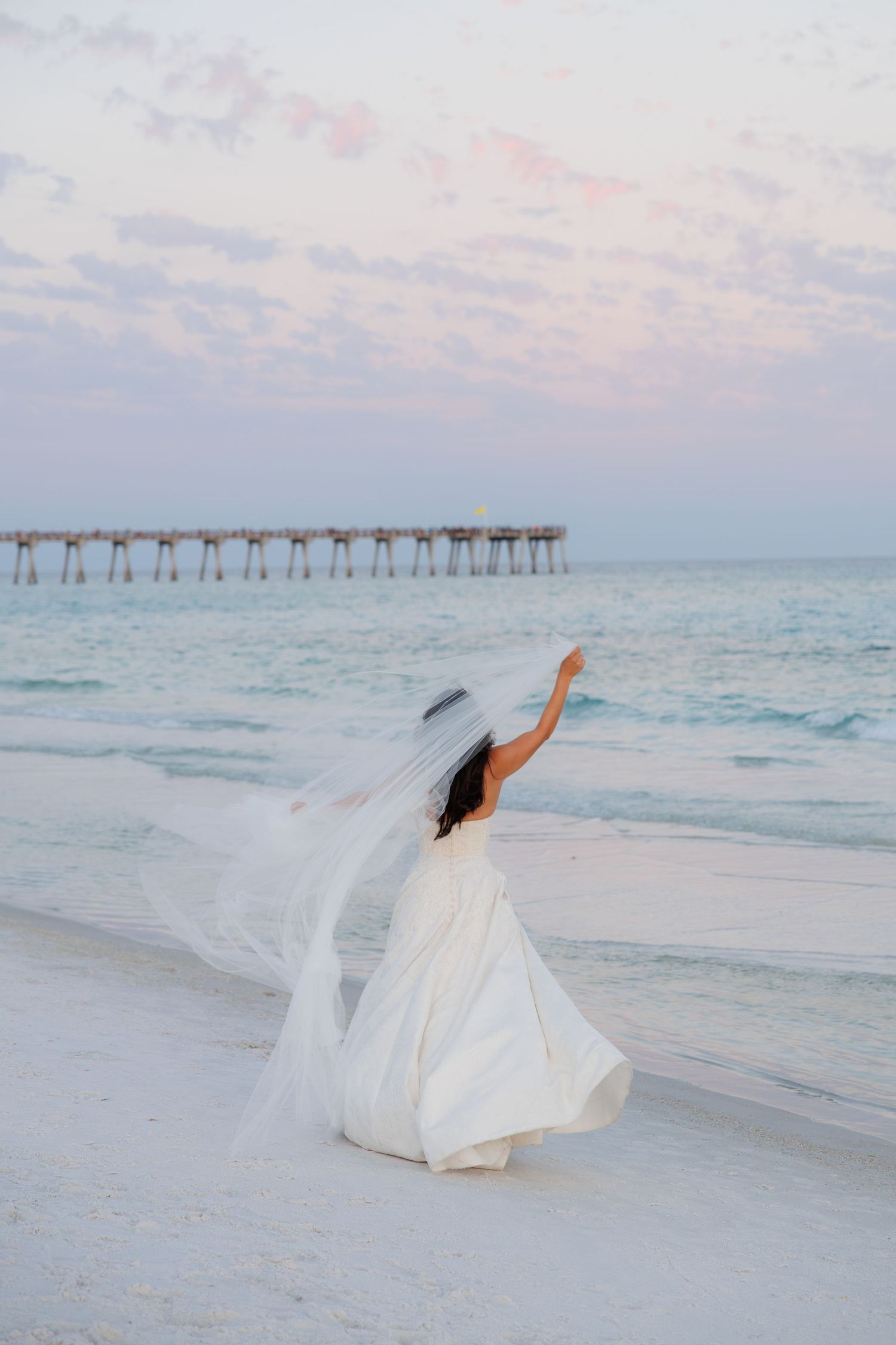 Bride in white gown and veil walks on a beach at sunset with a pier in the background.