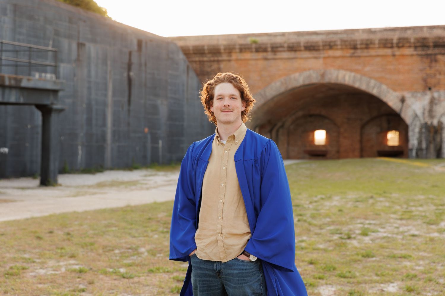 Person in blue coat stands smiling before historic brick fort archways on green lawn at golden hour.