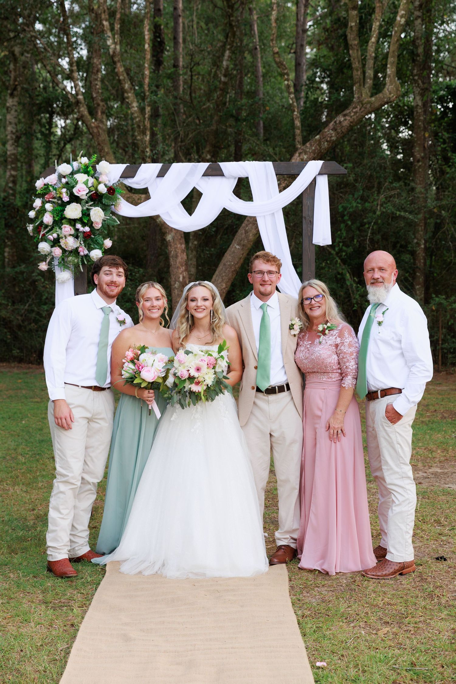 Wedding party in pink and mint green attire poses in front of white draped arch in outdoor woodland setting.