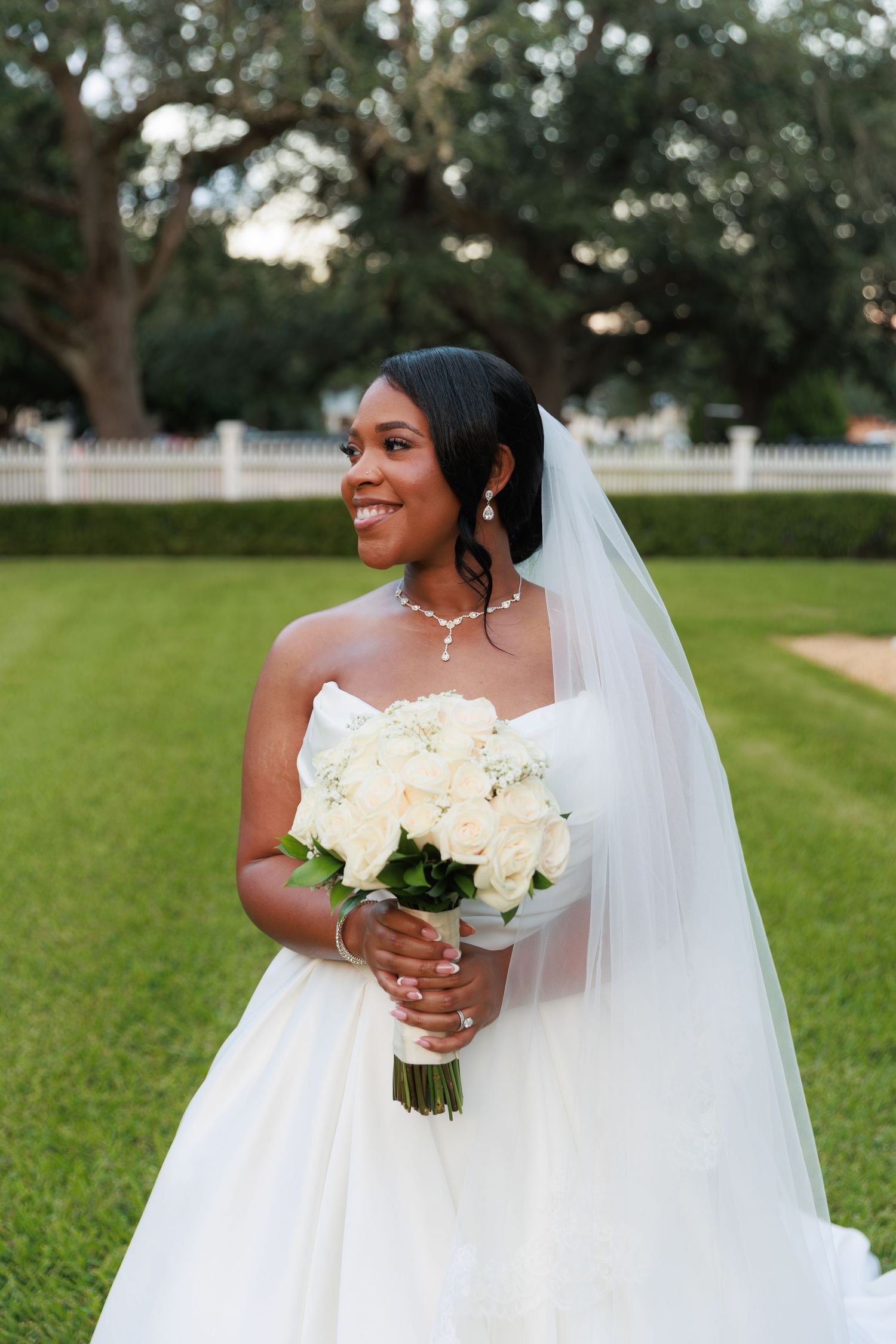 A bride in a strapless white wedding gown holds a bouquet of white roses while standing on a manicured lawn.