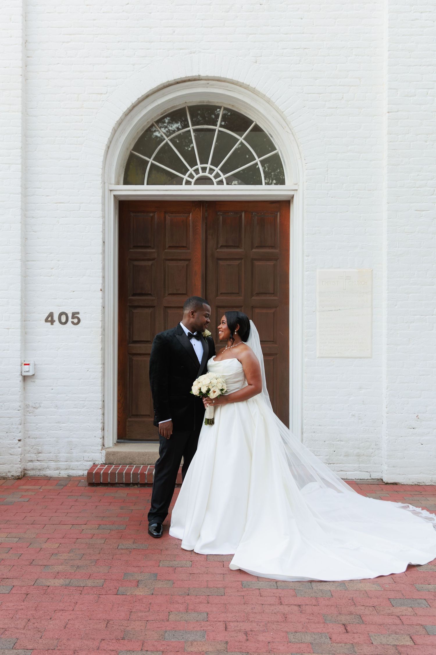 A couple shares an intimate moment in front of a historic white church entrance with a beautiful arched doorway.