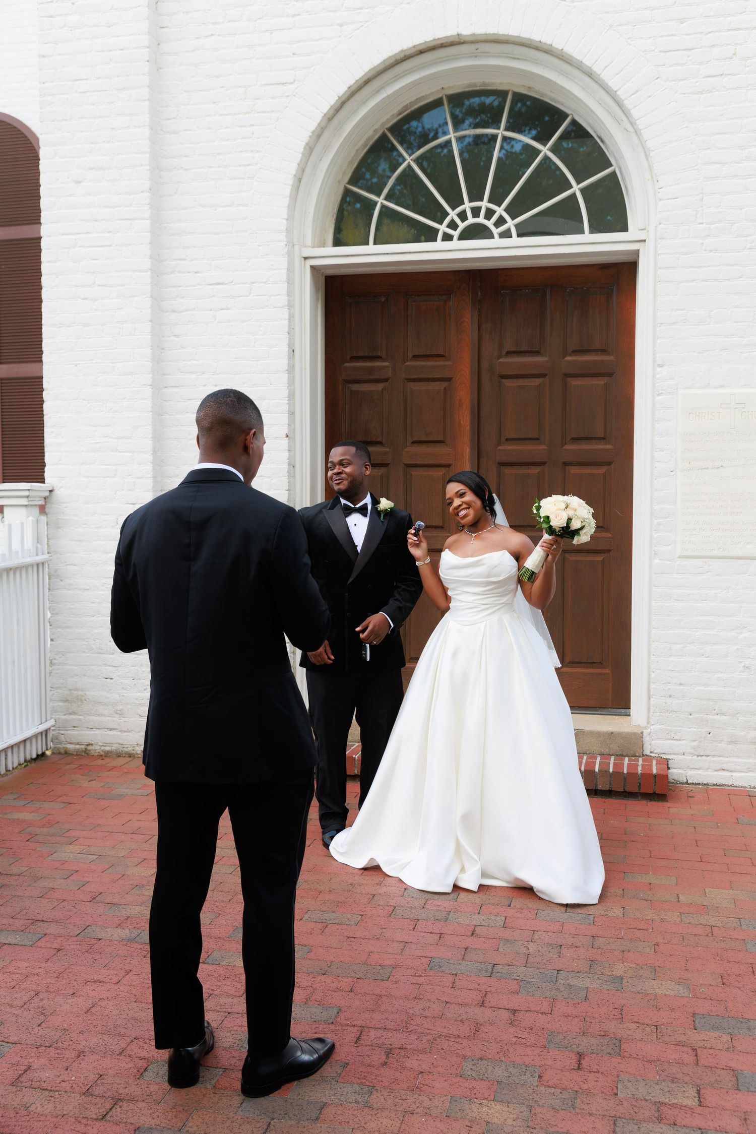 Wedding party in formal attire stands on red brick patio outside white church building with arched wooden doors.