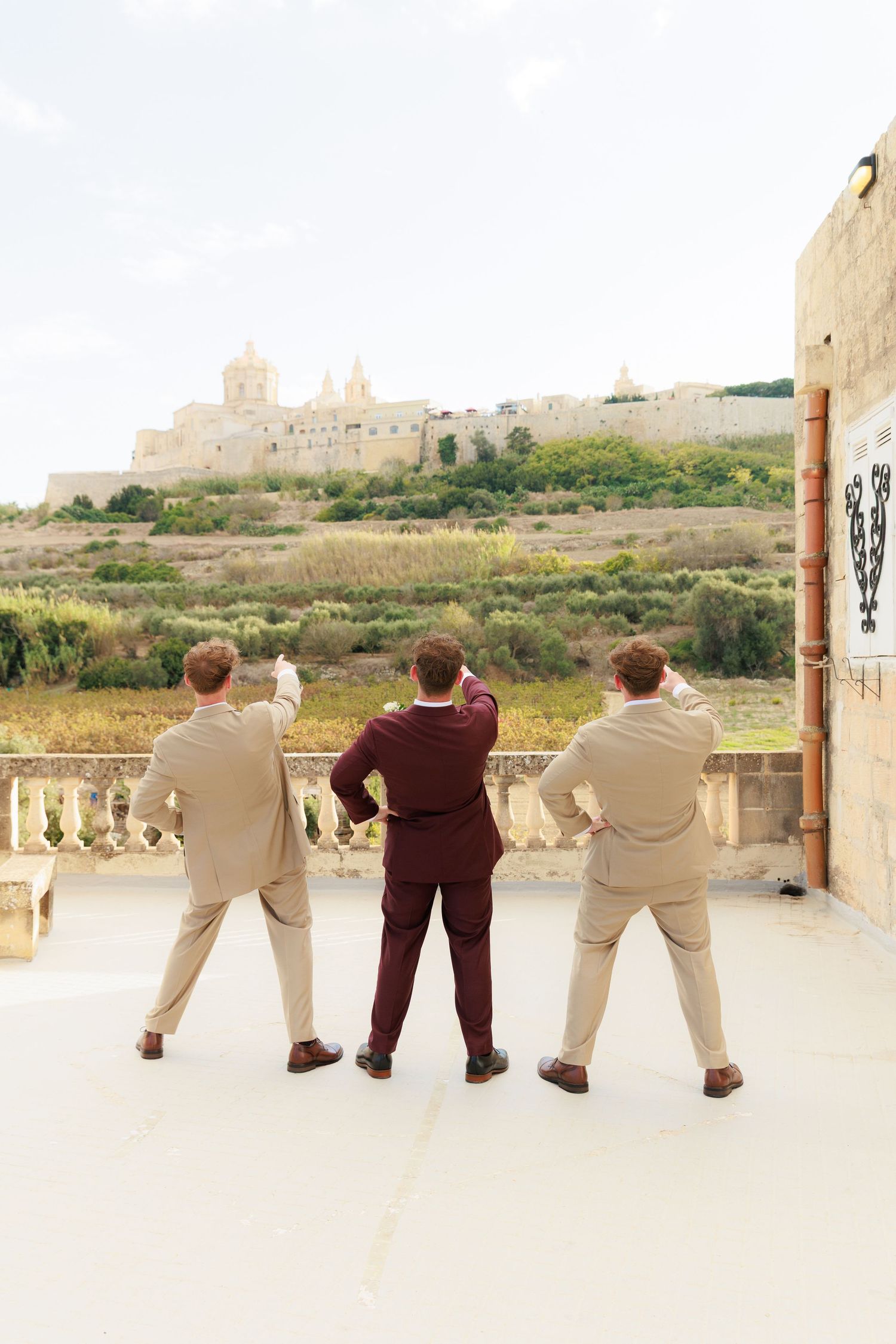 Three people in formal attire stand on a terrace overlooking a distant medieval fortress on a hilltop in Malta.
