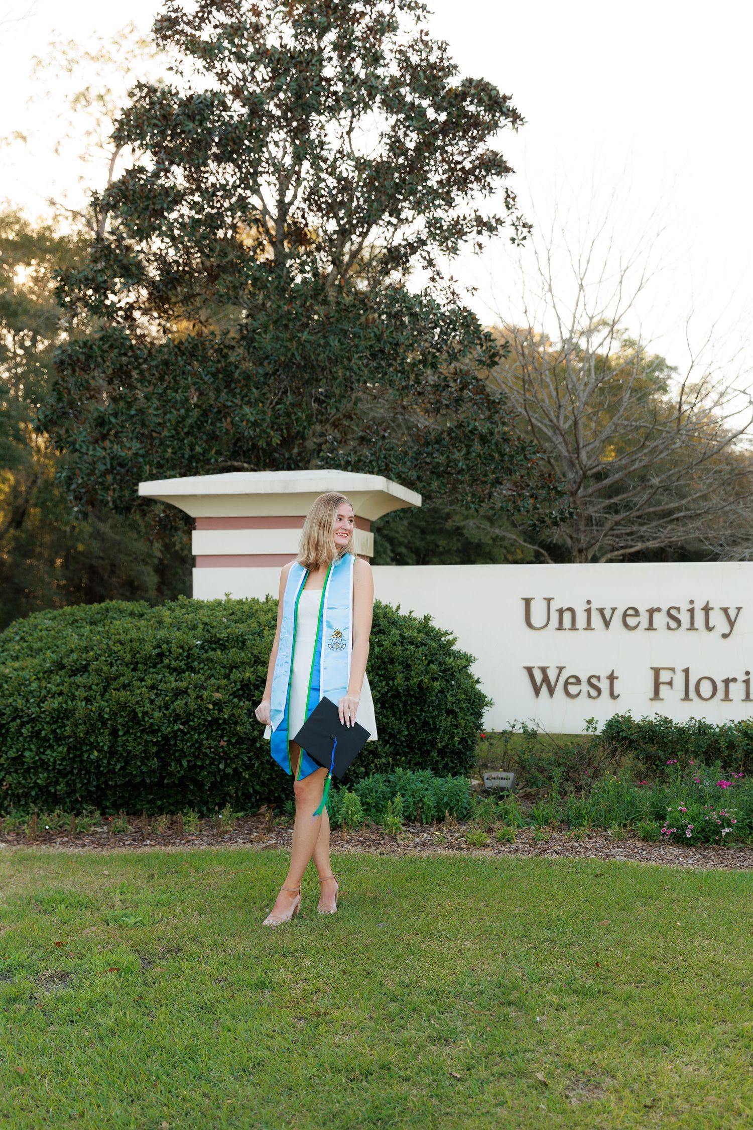 A graduate in cap and gown poses by the University of West Florida entrance sign during a sunny day.