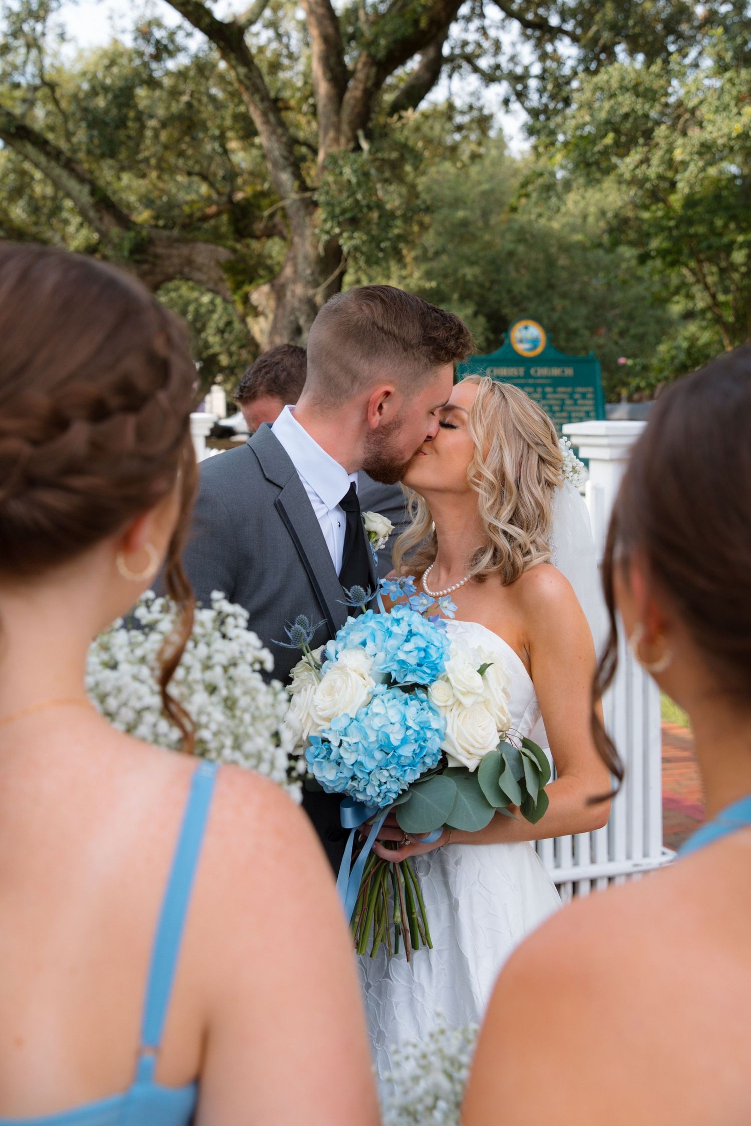 Bridesmaids watch as the happy couple shares a kiss outdoors with blue hydrangea bouquets.