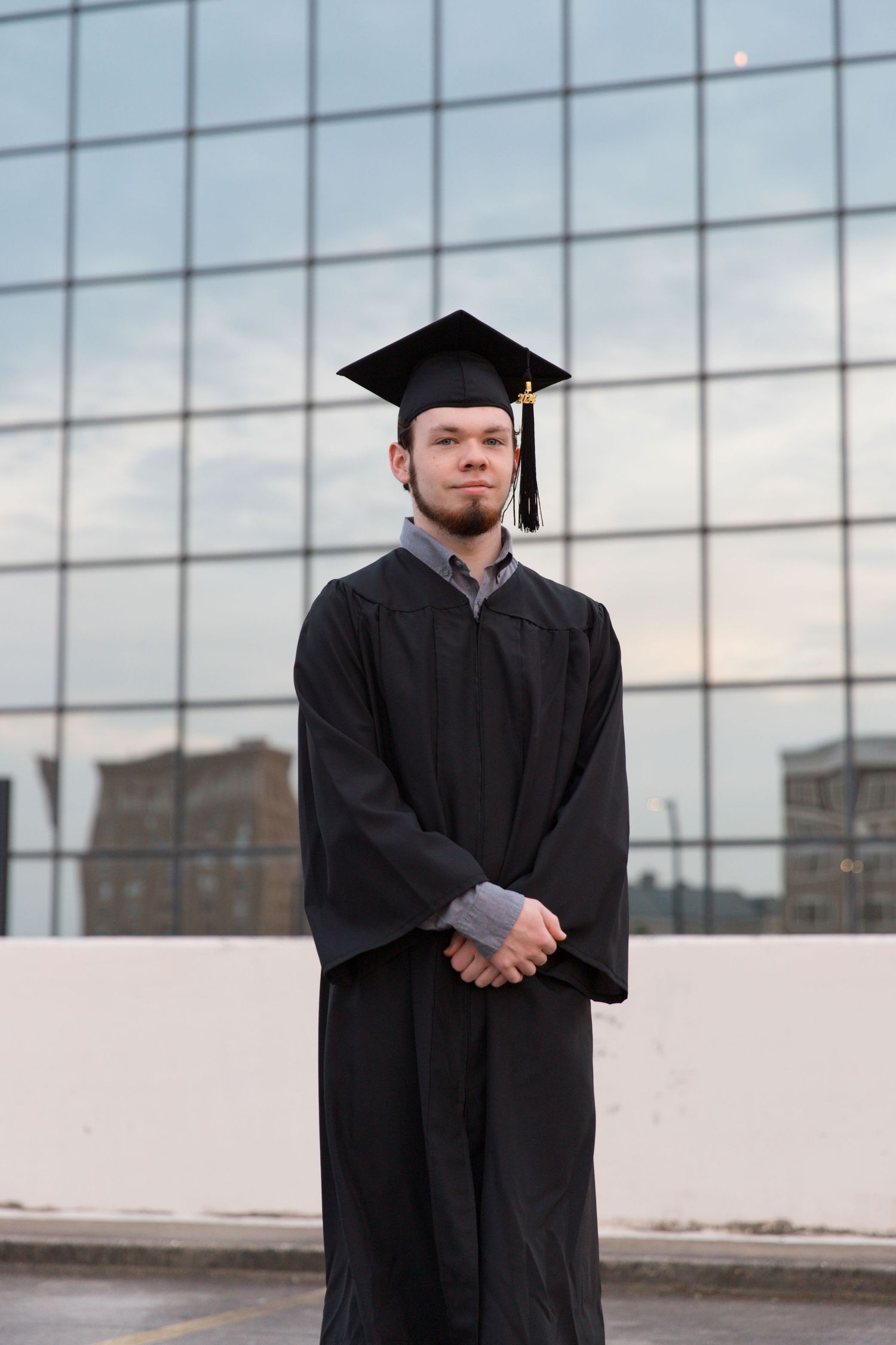 Graduate in traditional black graduation attire posed formally against glass building backdrop.