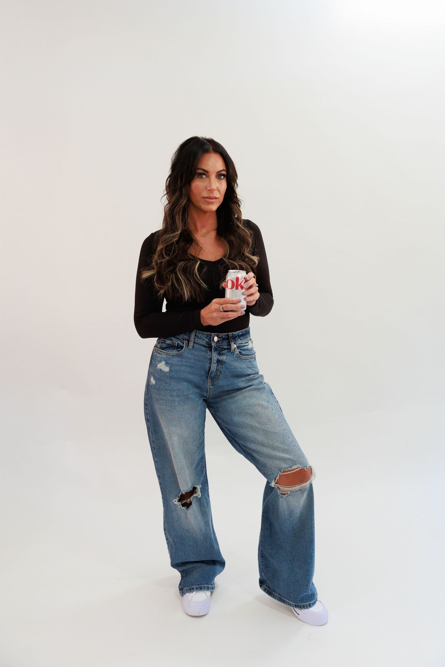 Woman in black top and distressed wide-leg jeans poses against white background.