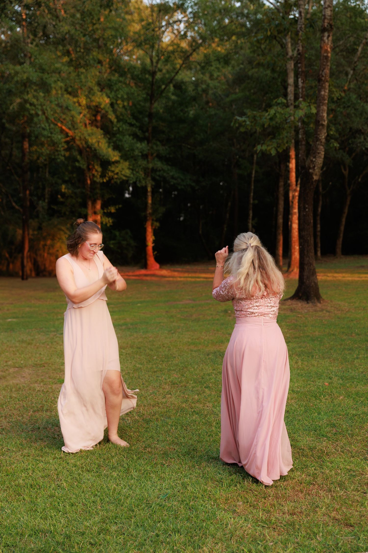 Two people in pink and blush dresses dance joyfully in a grassy park during golden hour.
