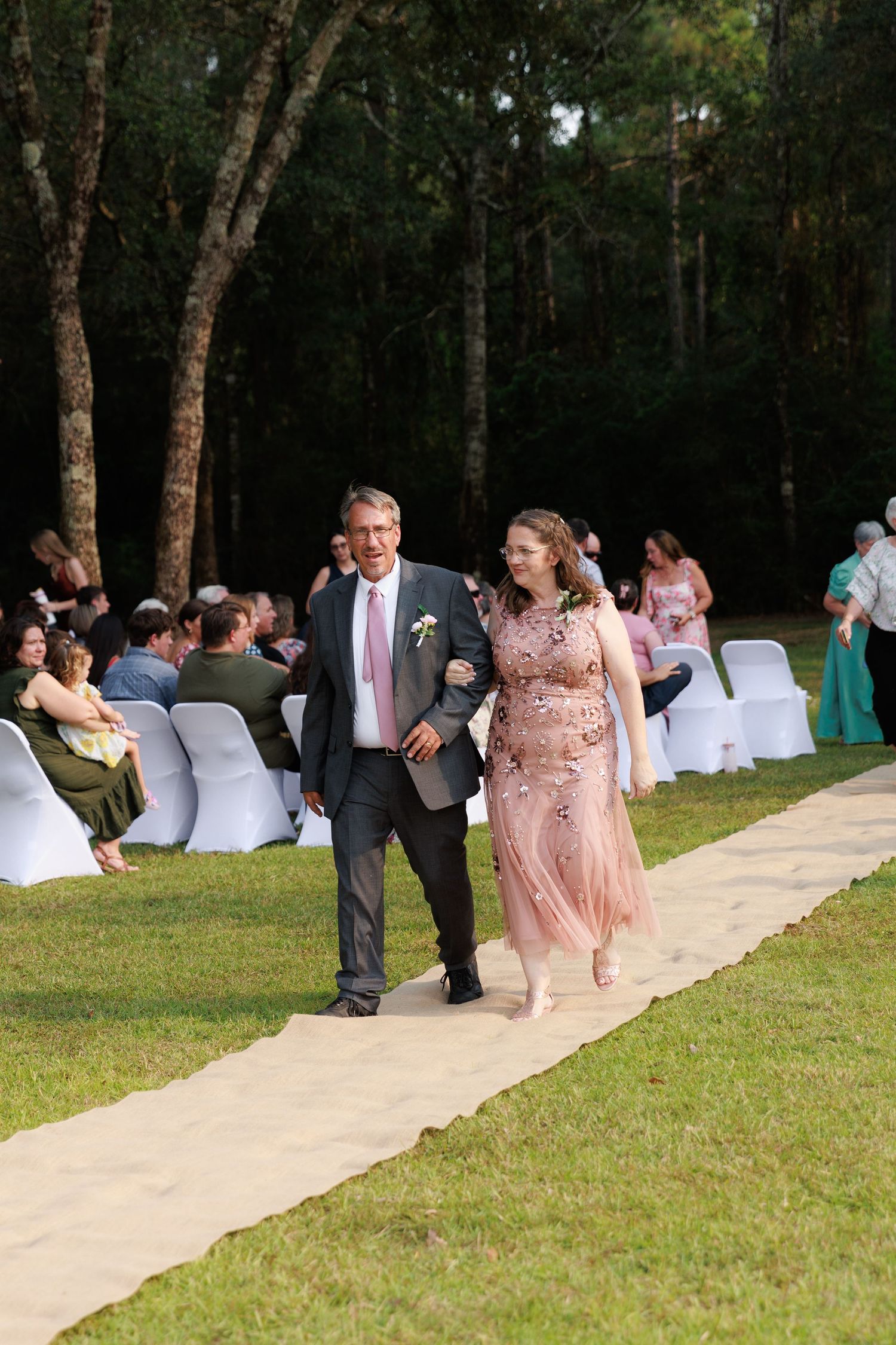 Wedding guests seated in white chairs along a tan aisle runner during an outdoor forest ceremony.
