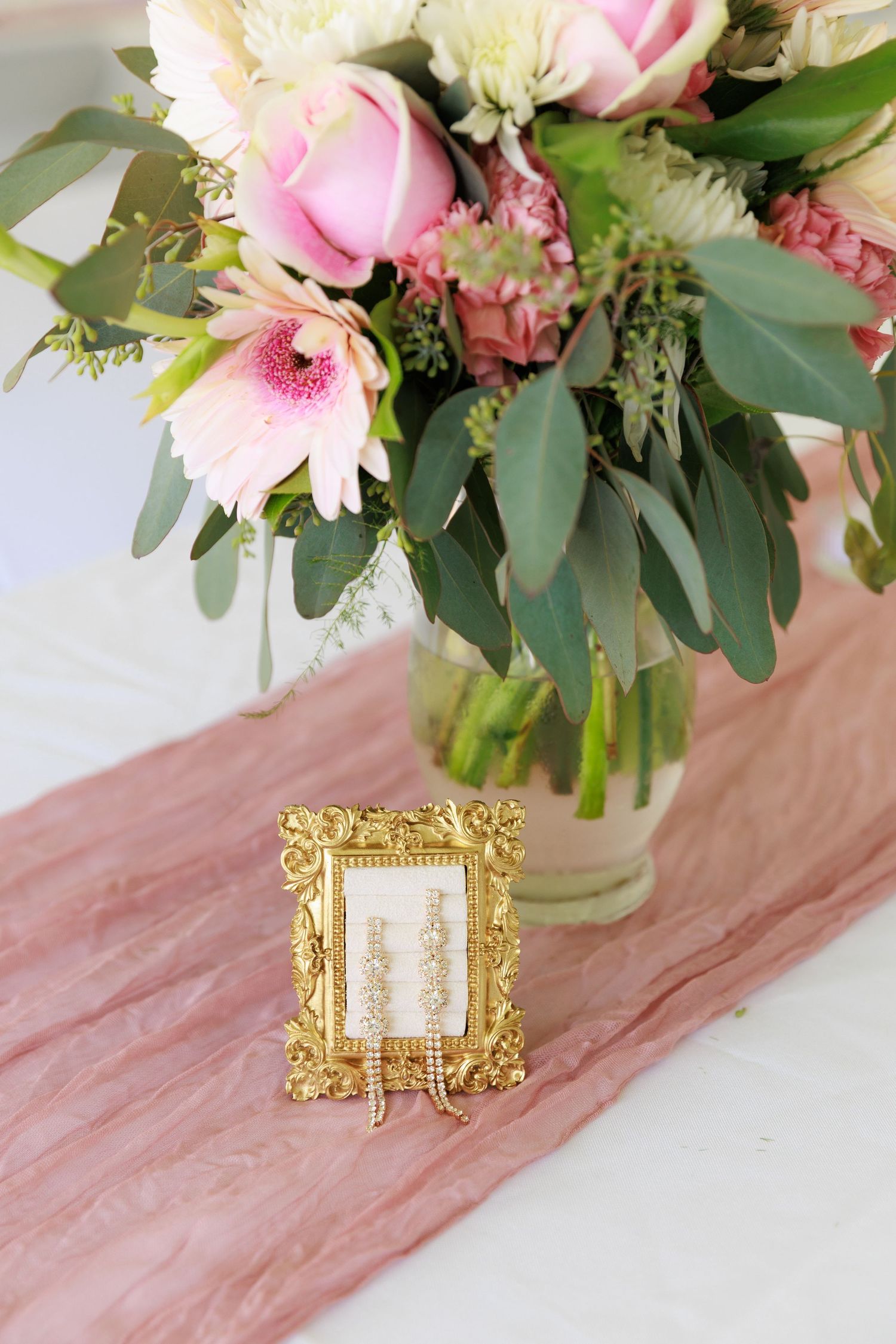 Pink and white floral arrangement in a glass vase with eucalyptus leaves and ornate gold frame on pink table runner.