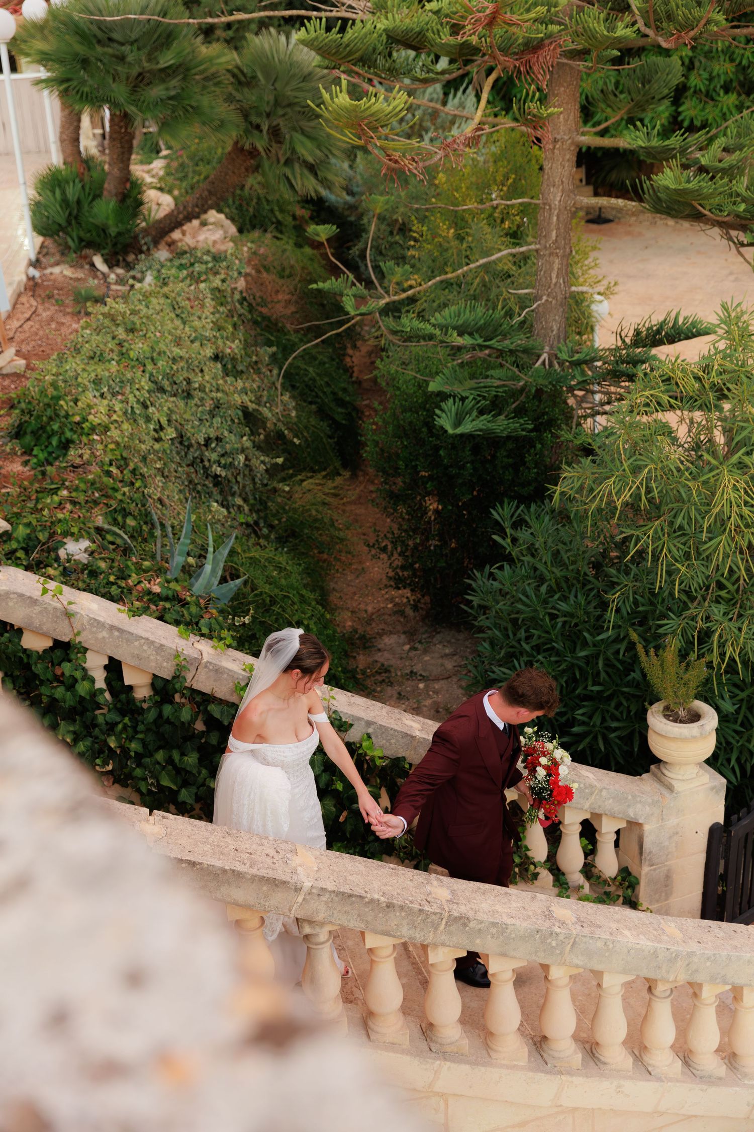 A couple descends stone garden stairs surrounded by lush greenery and palm trees at an elegant outdoor venue.