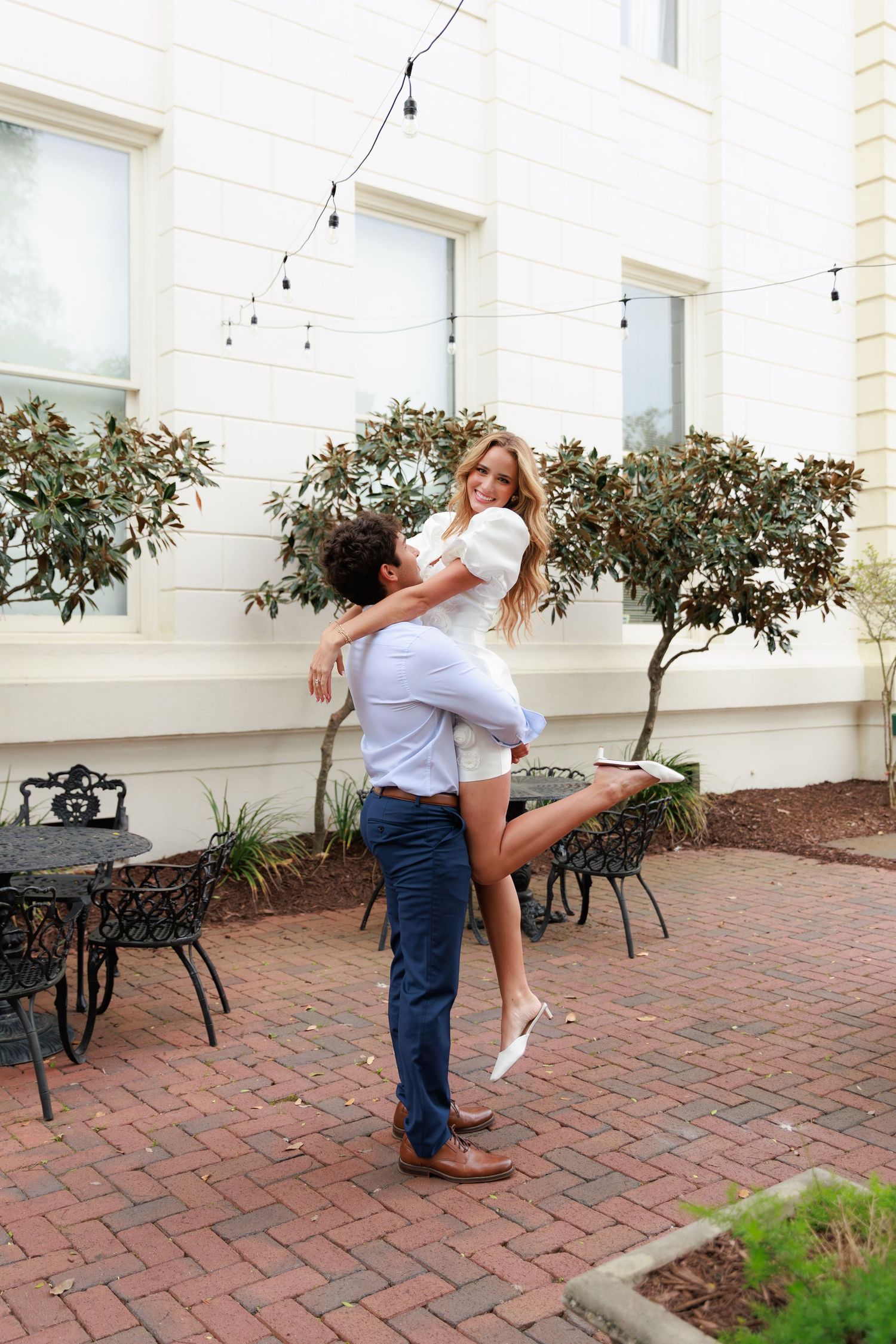 A romantic couple embraces on a brick patio with string lights and shrubs in the background.