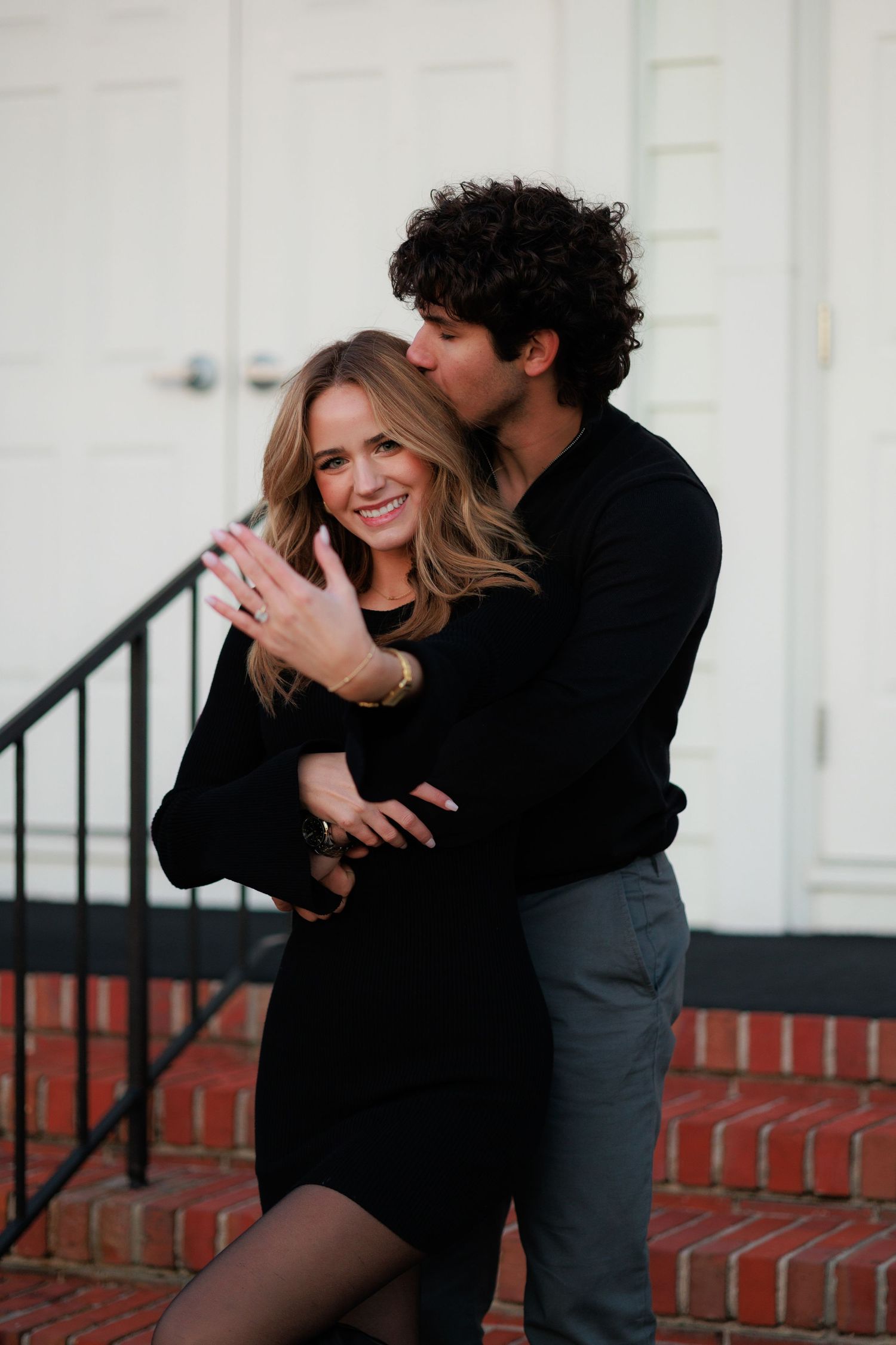 A couple embraces and laughs together on brick steps in front of a white building.