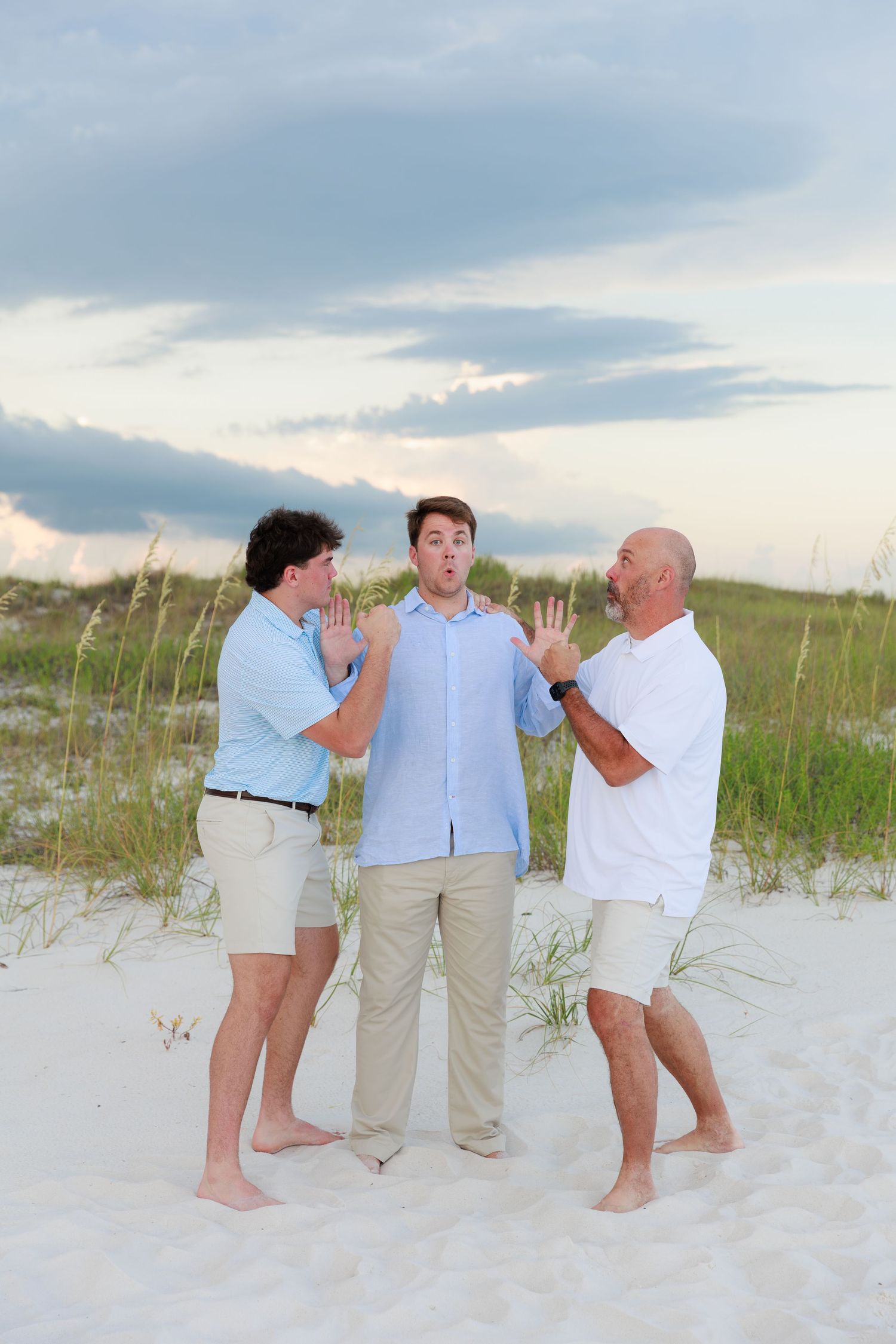 Three people in casual beach attire playfully interact on white sand dunes at sunset.