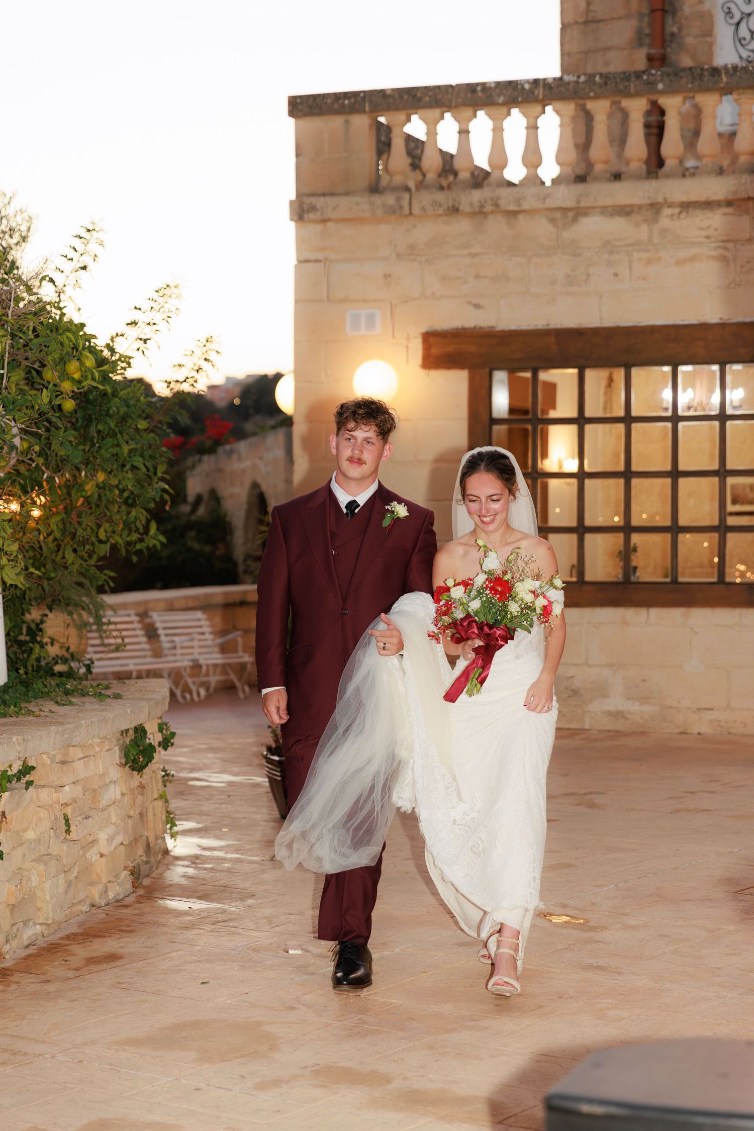 A couple poses for wedding photos on a stone patio with red flowers and architectural details in the background.