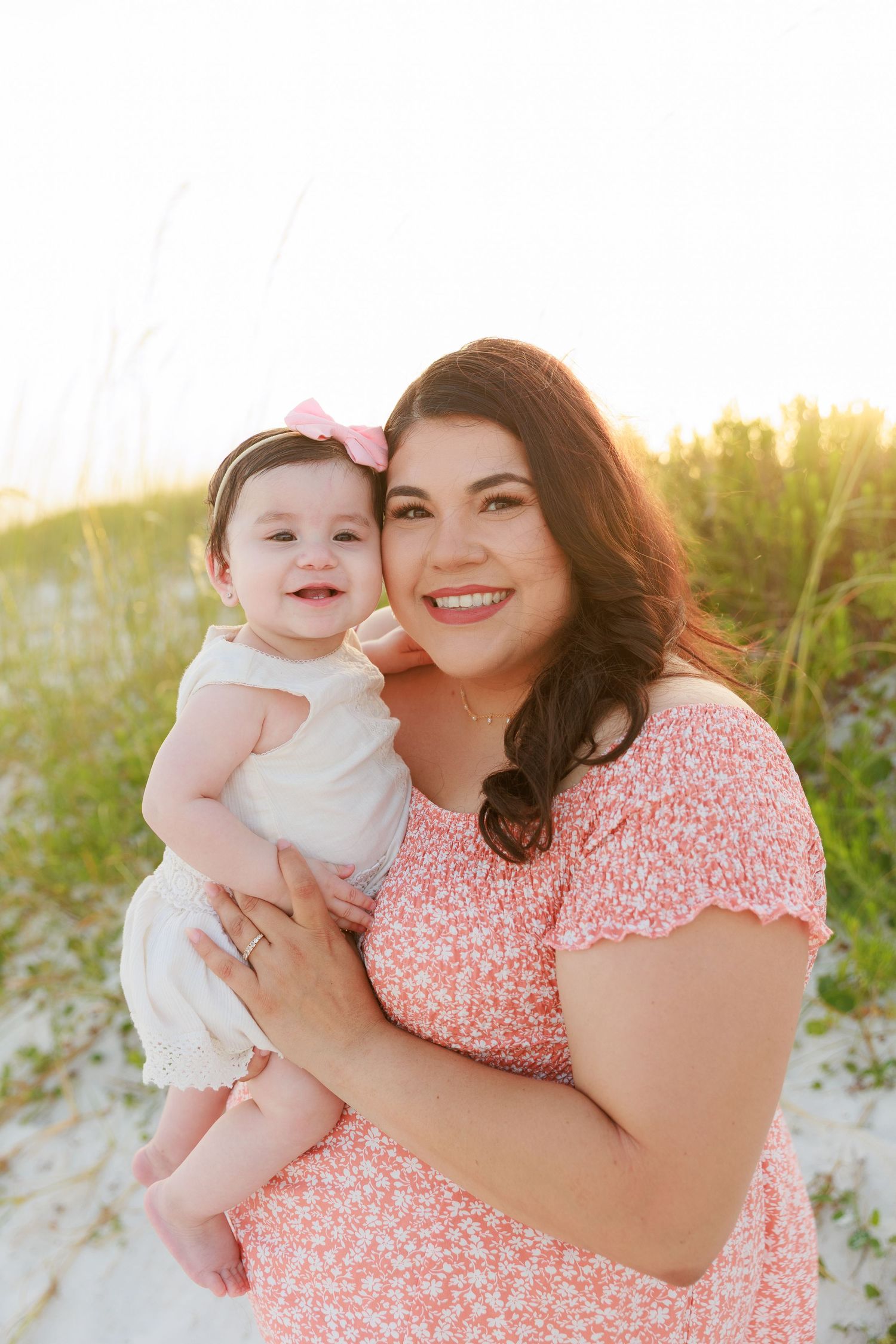 A smiling woman in a pink floral dress holds a happy baby girl with a pink bow at a sunny beach.