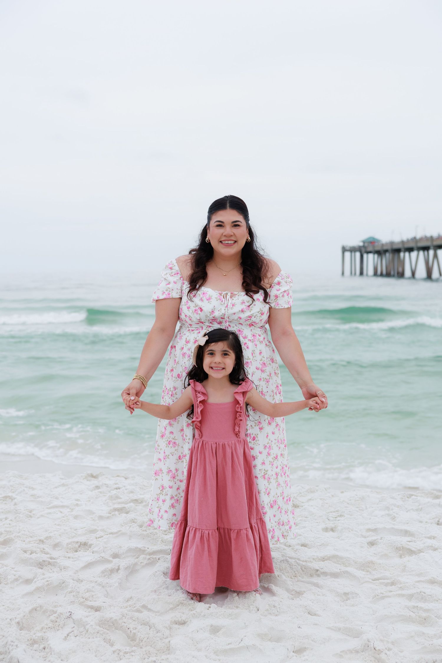 Mother in floral dress holding hands with young daughter in pink maxi dress on sandy beach near pier.