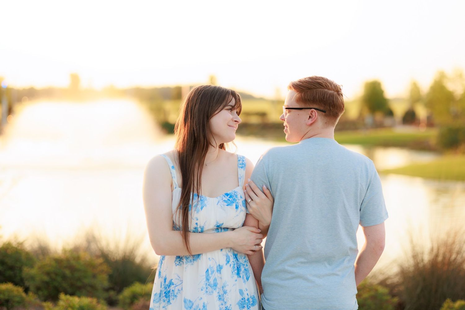 Couple sharing a romantic moment by a sunlit lake, woman in floral dress and man in blue shirt during golden hour.