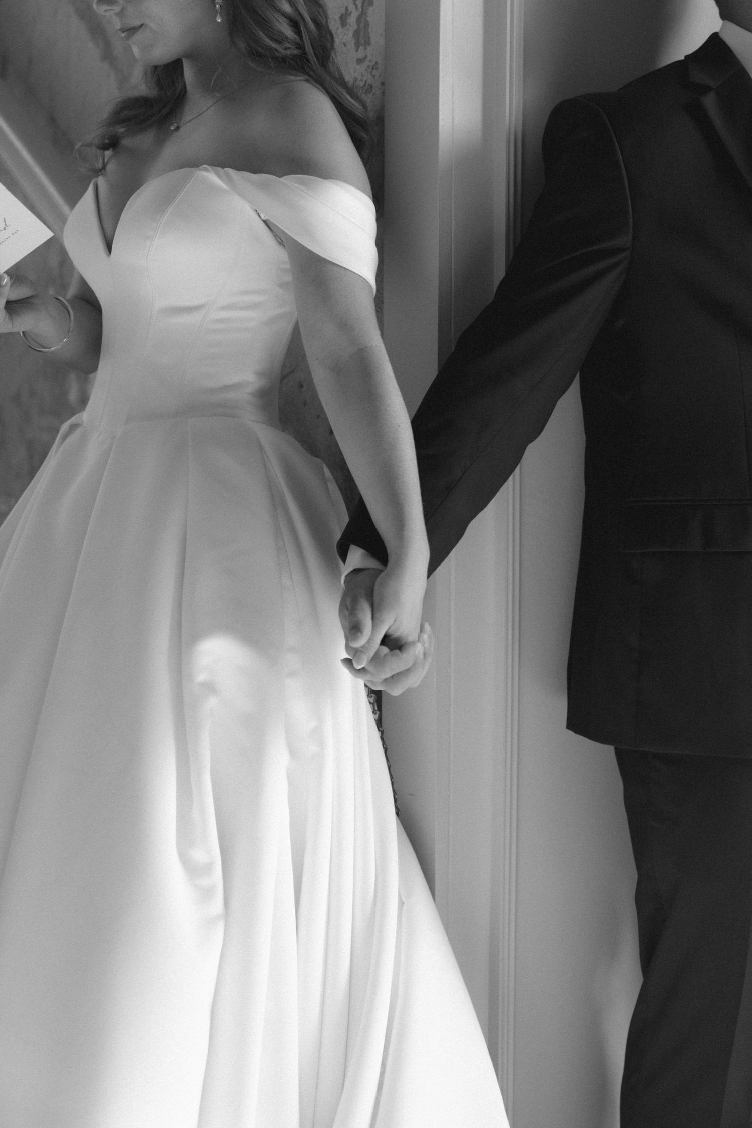 Black and white photo of bride in off-shoulder gown holding hands with groom in dark suit at wedding ceremony.