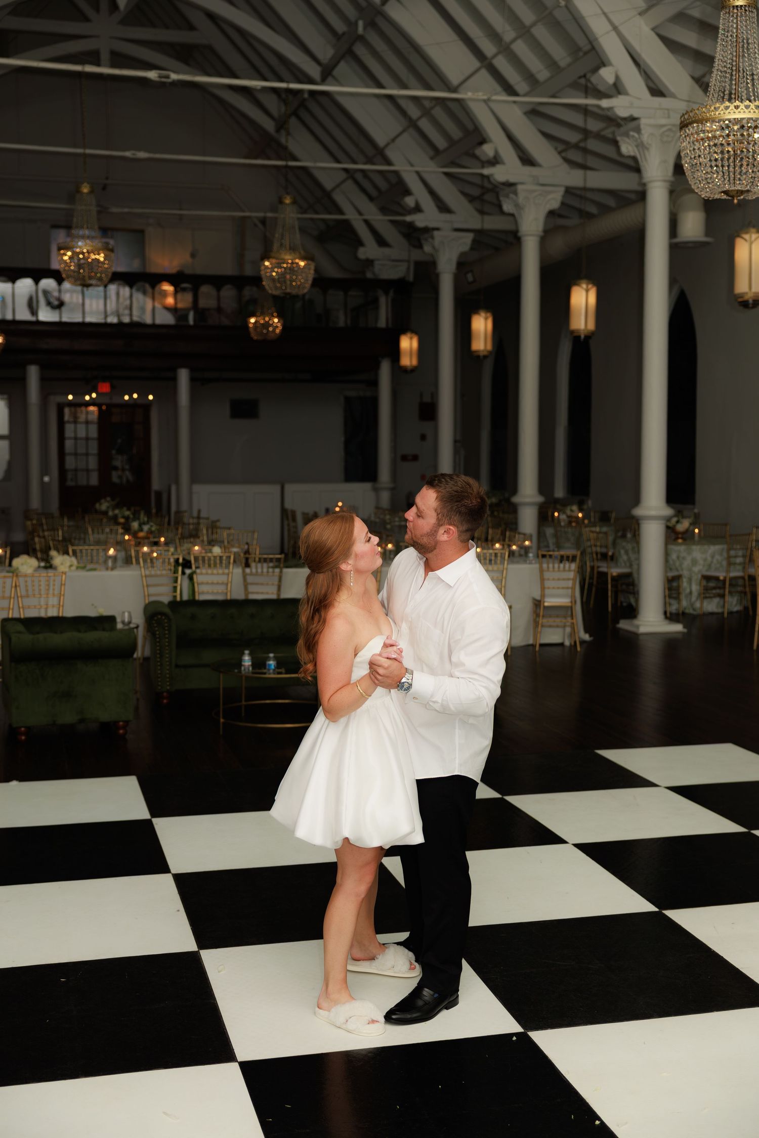 Newlywed couple shares romantic first dance on checkered floor in elegant chandelier-lit wedding venue.
