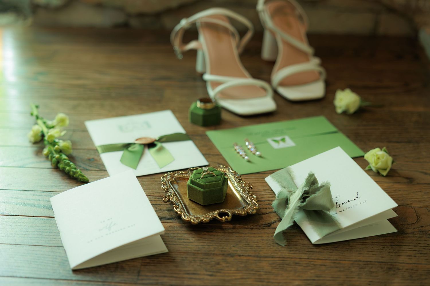 Elegant wedding flat lay with green stationery, velvet ring box, boutonniere, and white strappy heels on wood floor.