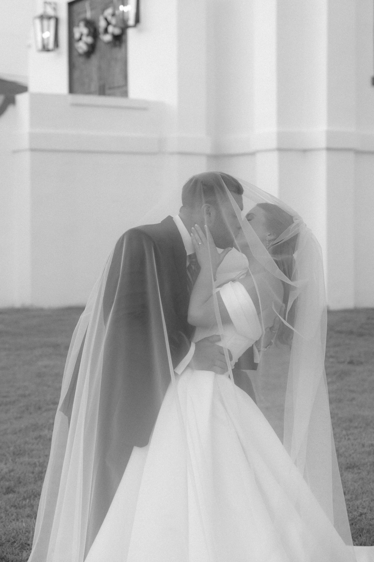 Black and white photo of bride and groom kissing beneath flowing veil outside white wedding venue.