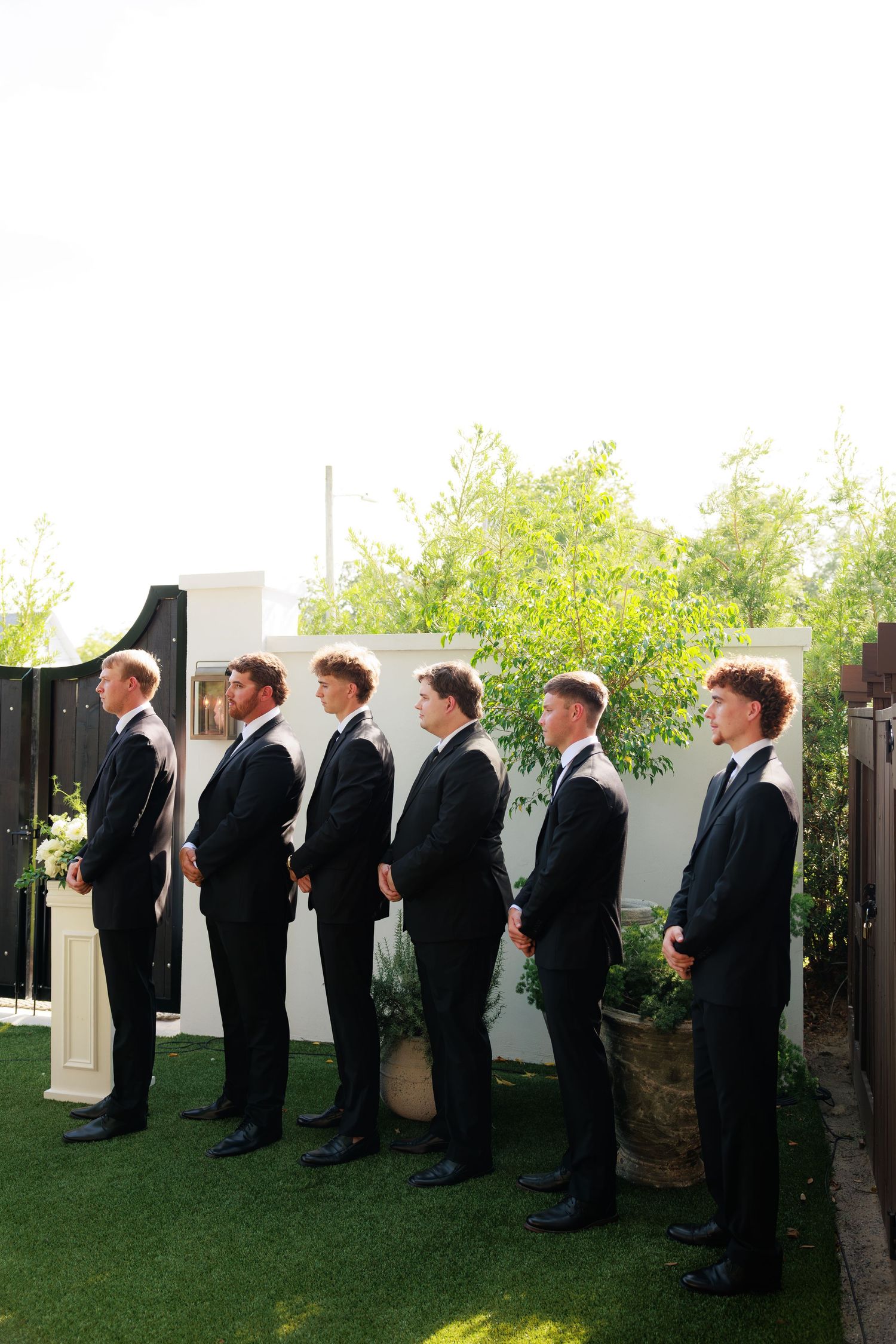 Six groomsmen in black tuxedos stand in a line at outdoor wedding.