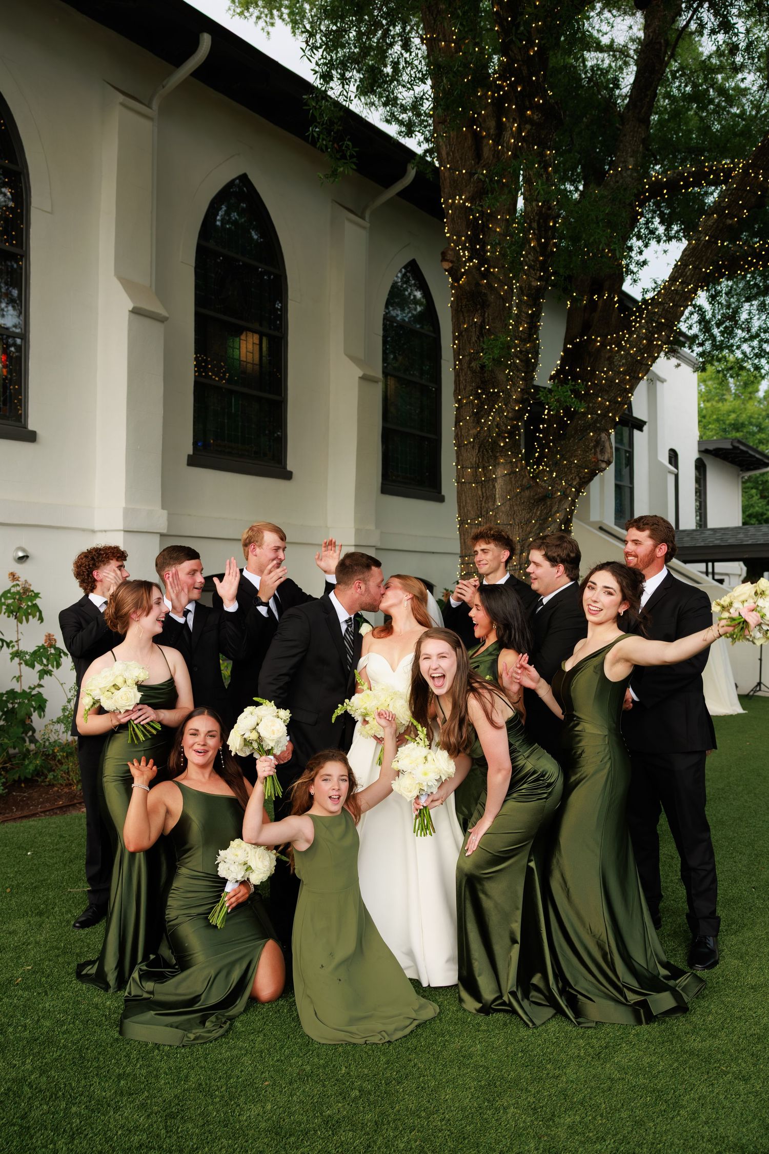 Wedding party with bridesmaids in green gowns and groomsmen in black suits celebrate as bride and groom kiss.