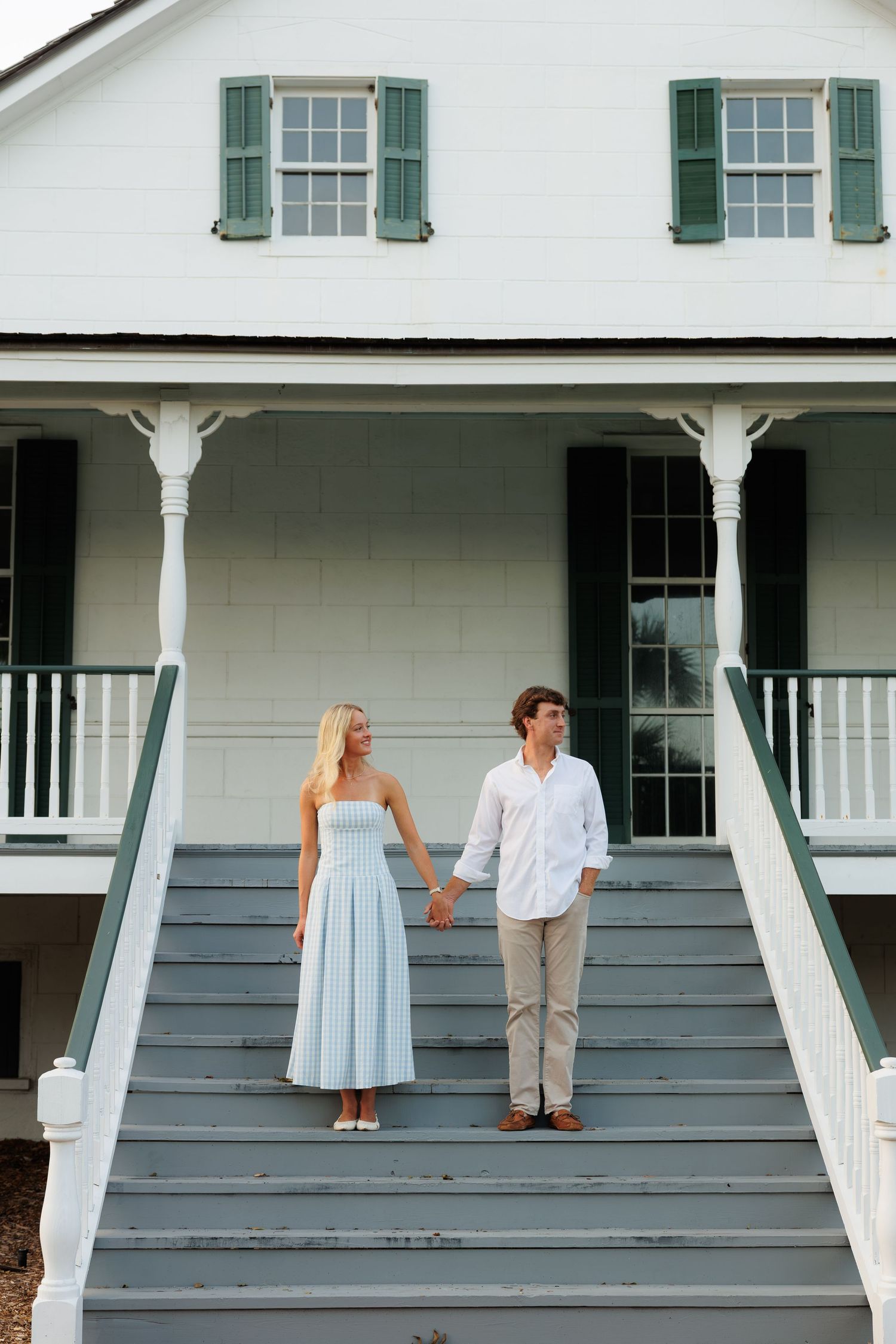 A couple holds hands on gray steps of a white colonial home with green shutters.