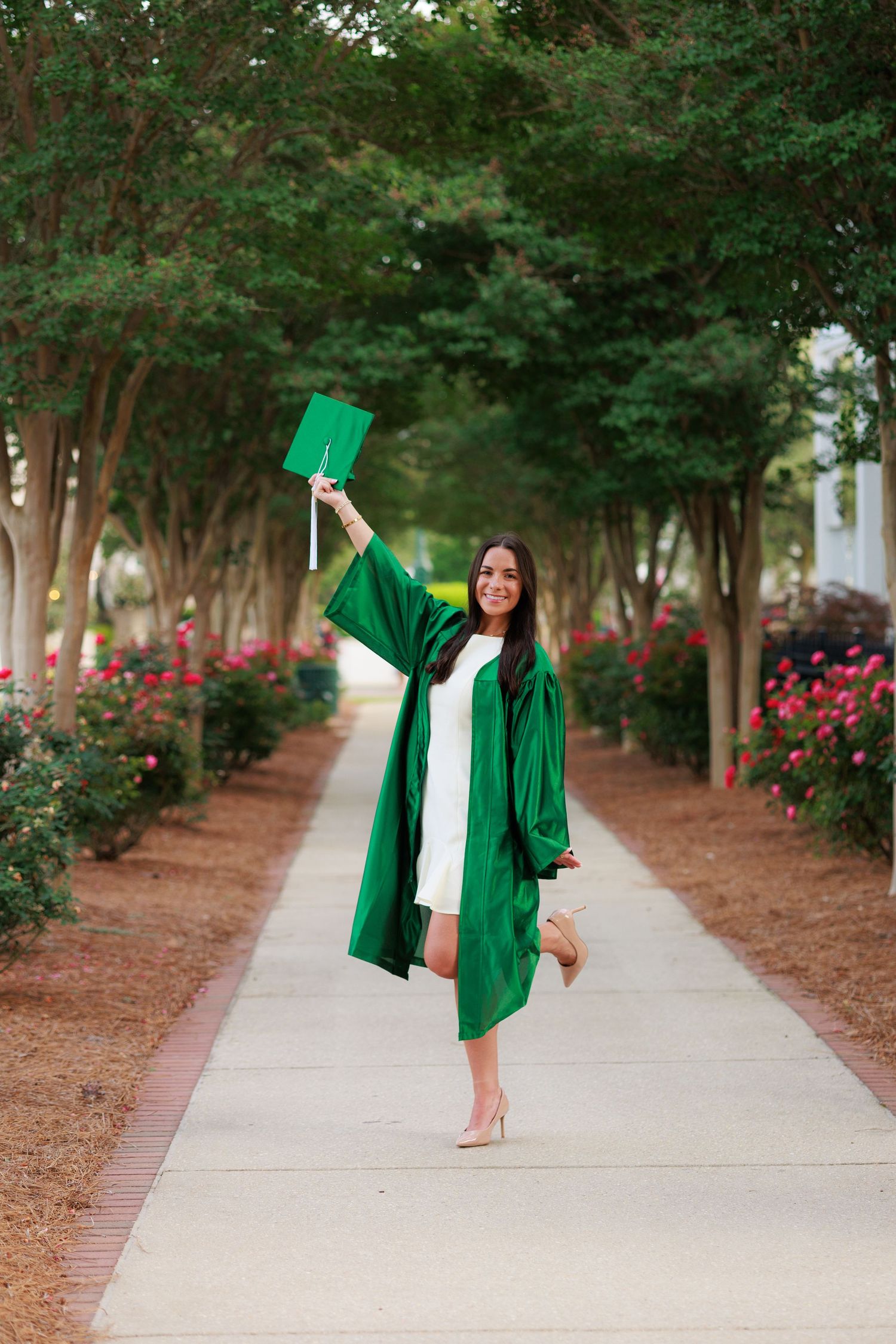 Young woman in green graduation gown and cap celebrating on a tree-lined walkway with colorful flowers.