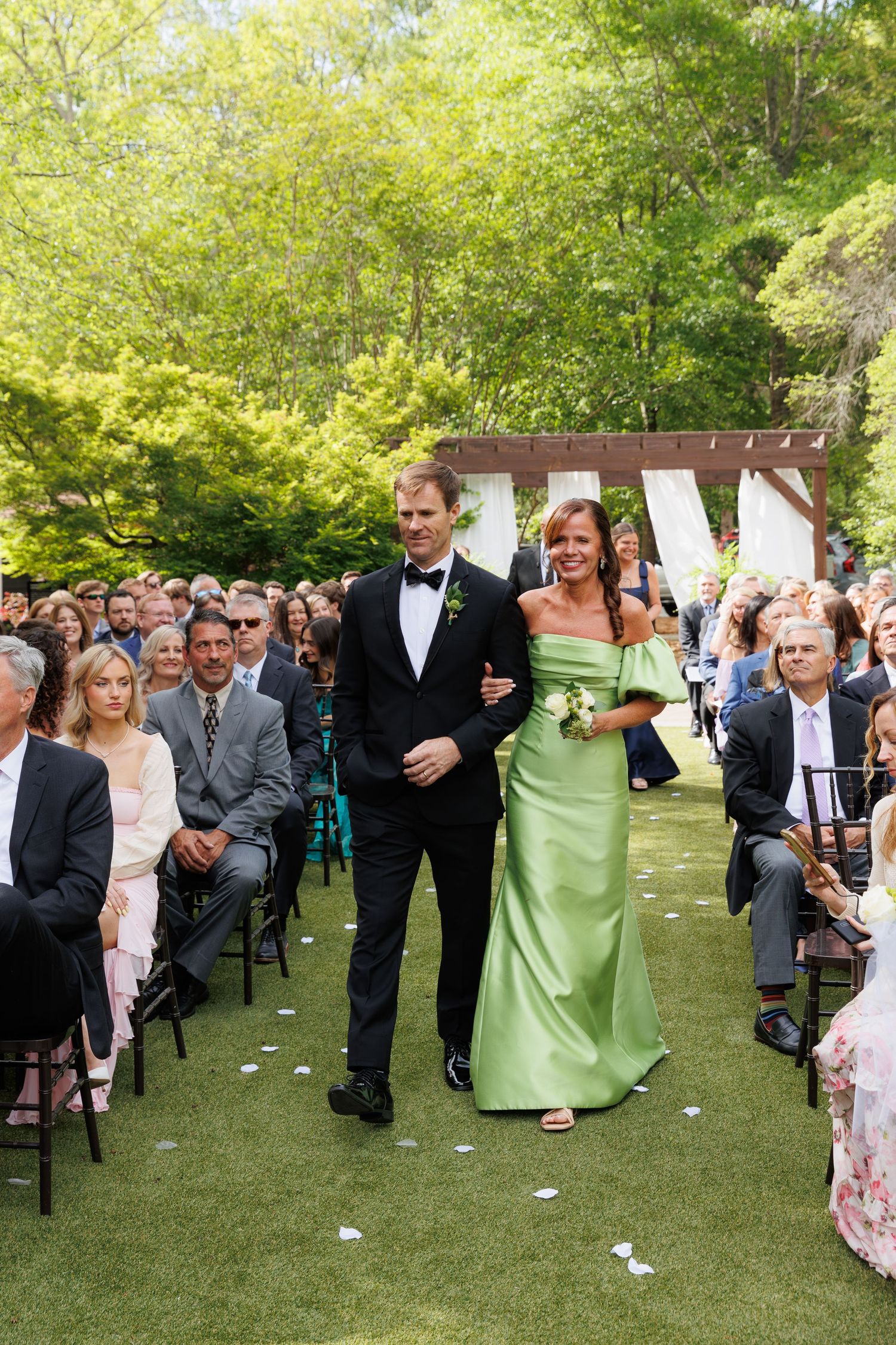 A man in a tuxedo escorts a woman in a green off-shoulder gown down an outdoor wedding aisle.